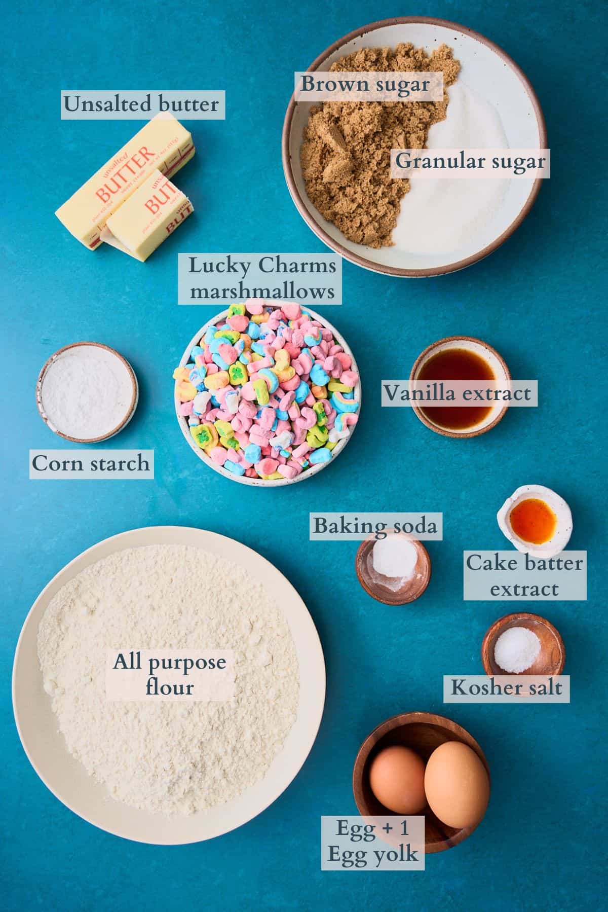 Ingredients to make Lucky Charms Cookies laid out on a deep green backdrop and labeled to denote each ingredient. 