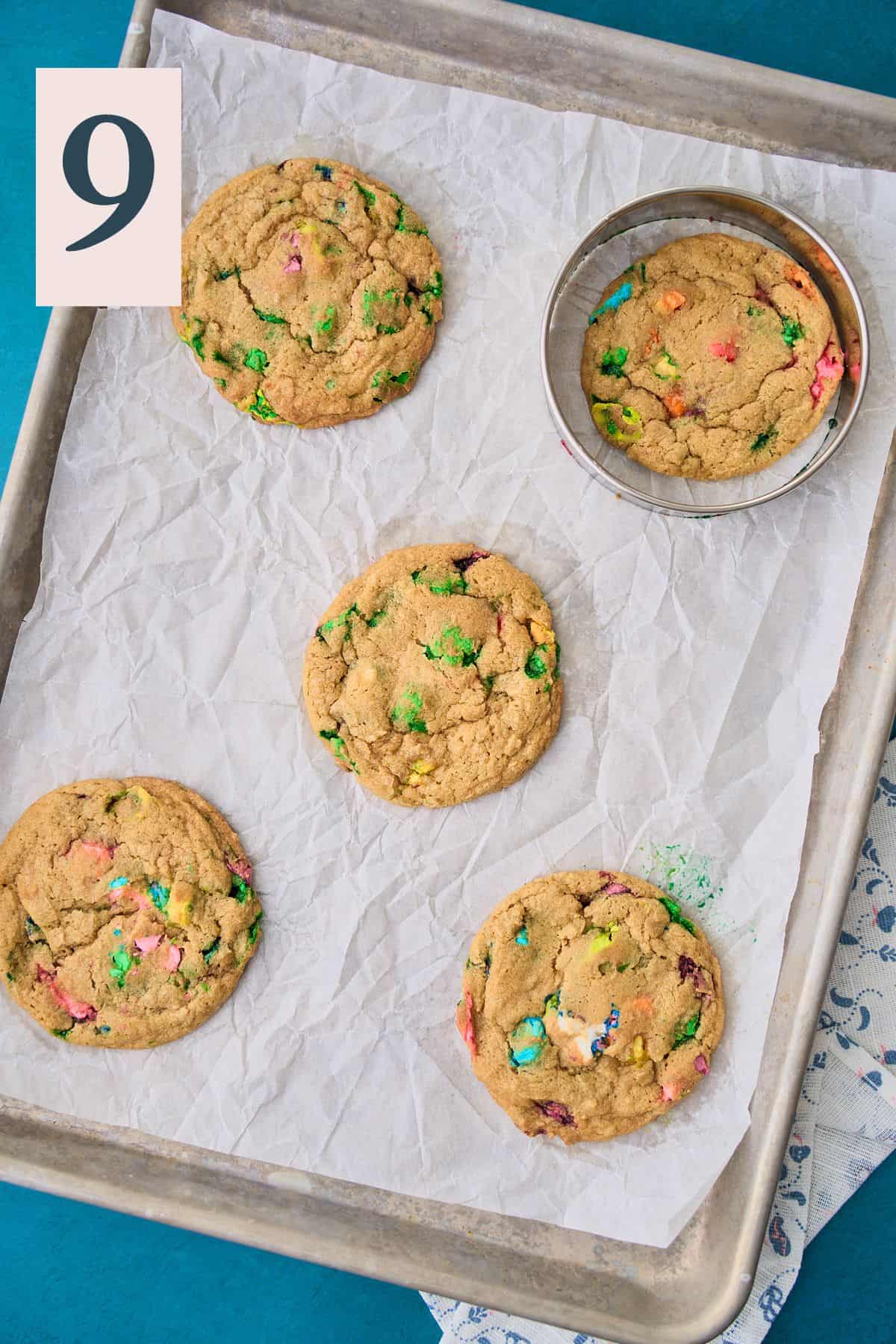 Baked lucky charms cookies on a parchment lined baking sheet with a cookie cutter around a cookie to scoot it into a round shape.