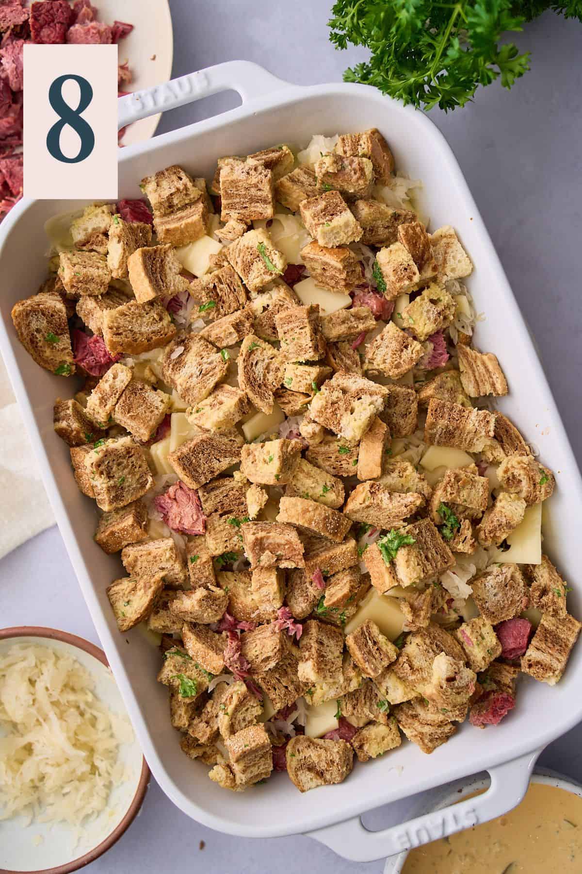 Bread pieces on top of corned beef, cheese and sauerkraut in a casserole dish. 