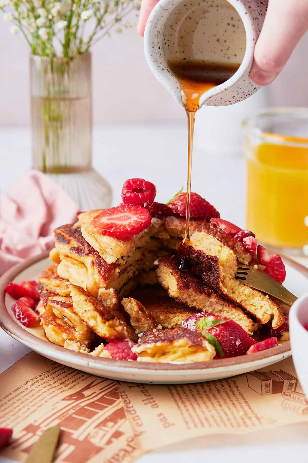 Stack of sweet cream pancakes with berries on top and a hand pouring maple syrup onto the stack, with a bite taken out of the side to reveal the delicious fluffy interior.