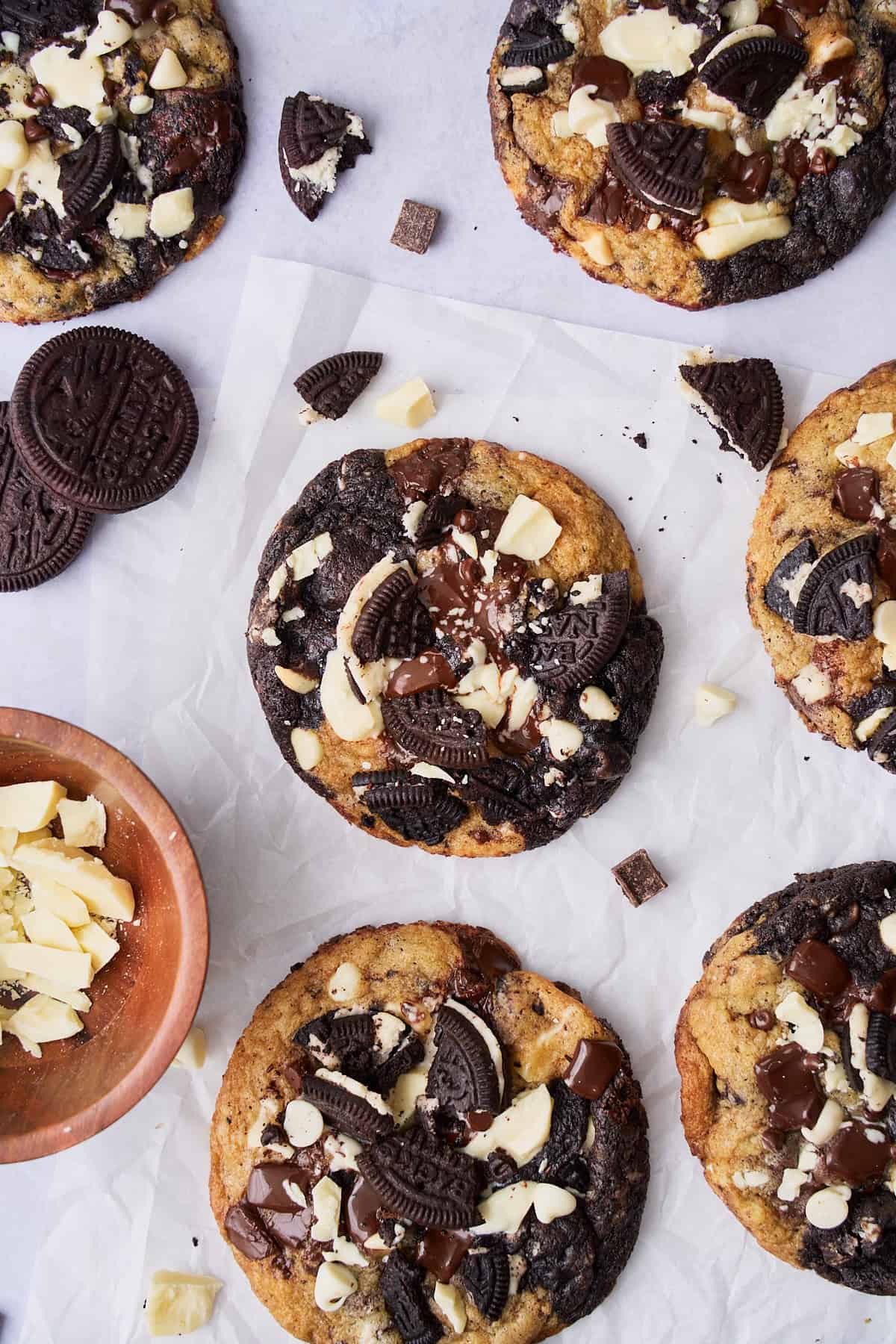 Marbled cookies and cream cookies on a wrinkled piece of parchment paper surrounded by pieces of white chocolate, and Oreo like sandwich cookies. 