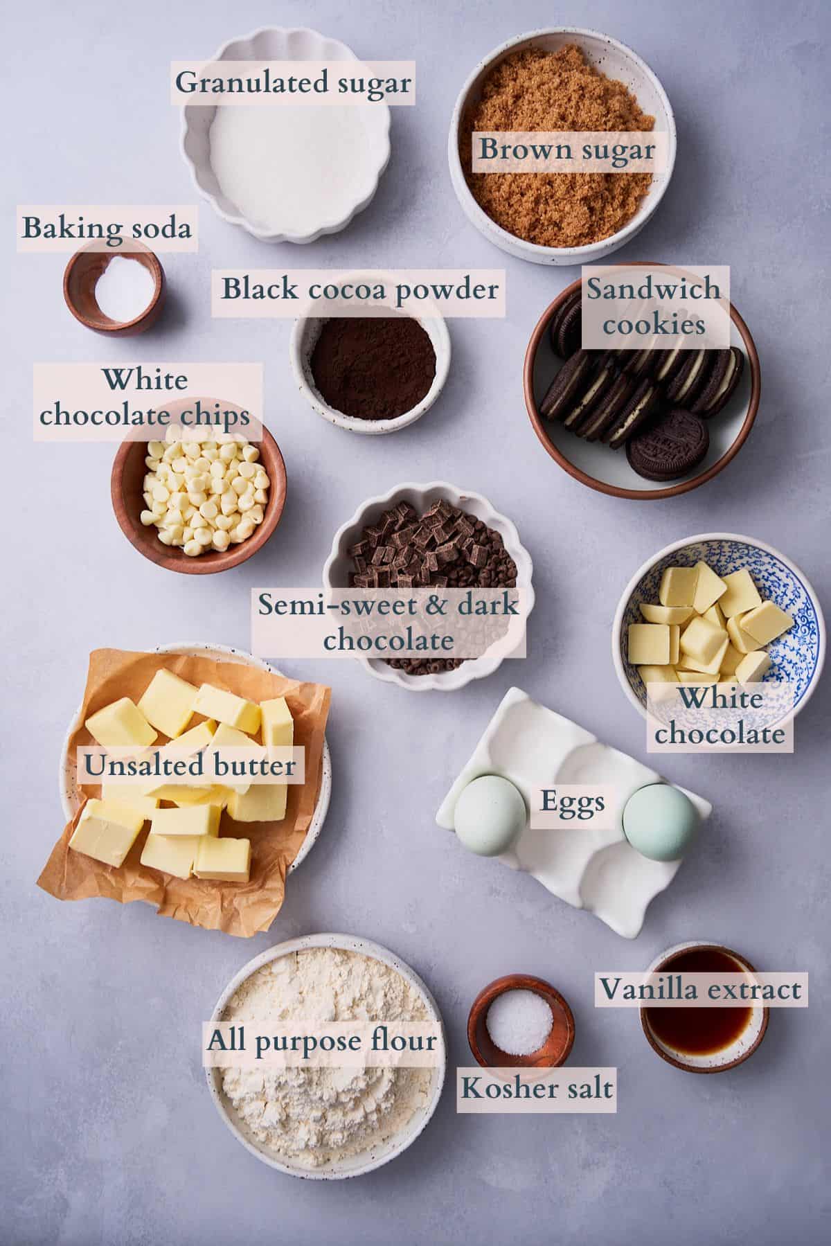 Ingredients to make cookies and cream cookies laid out on a table in small bowls and labeled to denote each ingredient.