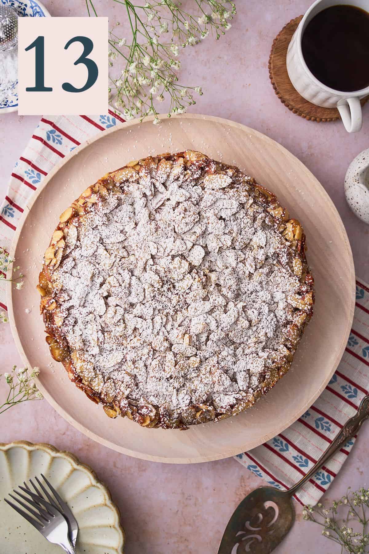 Swedish almond cake topped with a thin layer of powdered sugar, on a wooden plate, surrounded by white flowers, and coffee. 