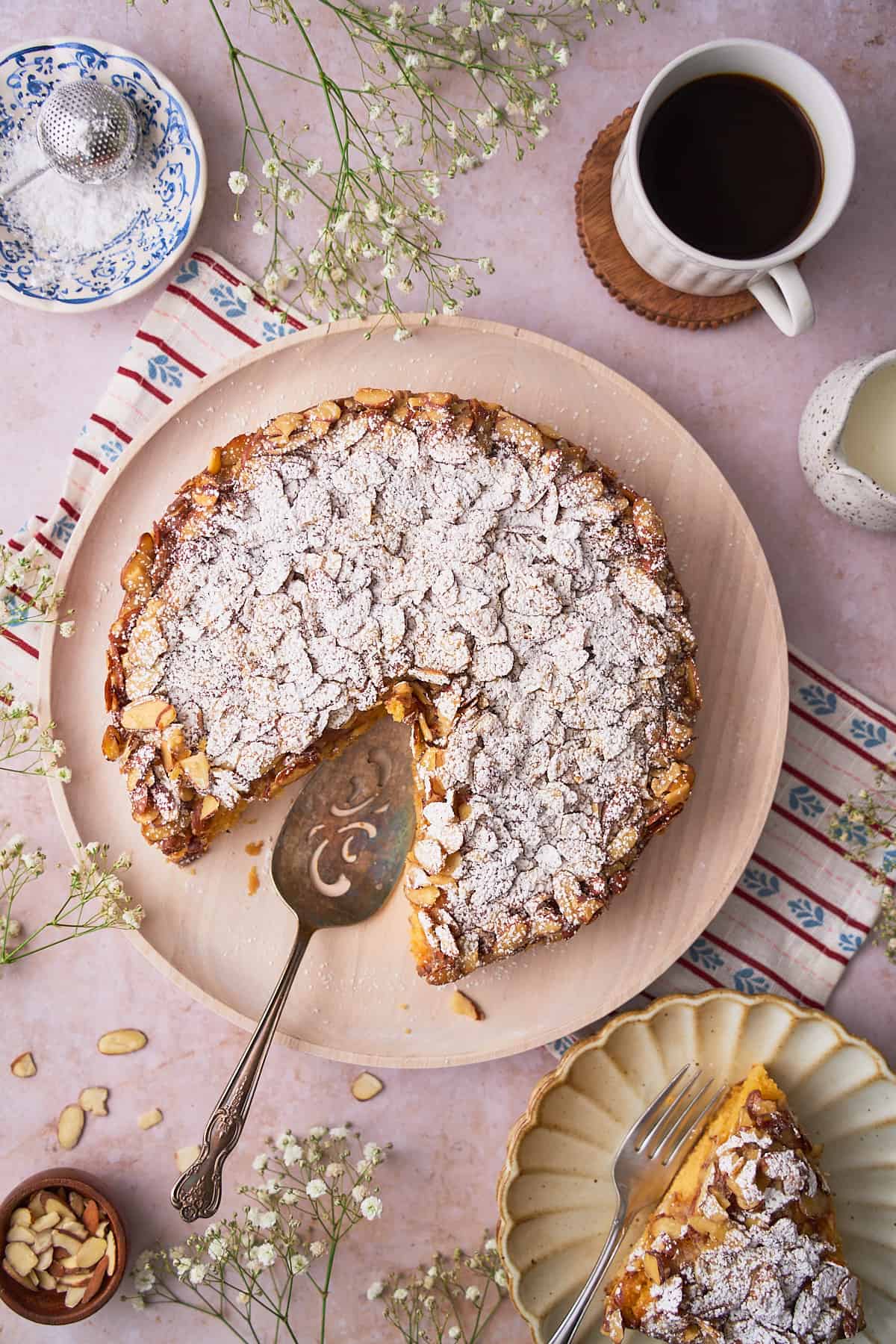 Overhead view of a Swedish almond cake on a wooden serving plate, with a slice cut out, and surrounded by white flowers, and coffee. 