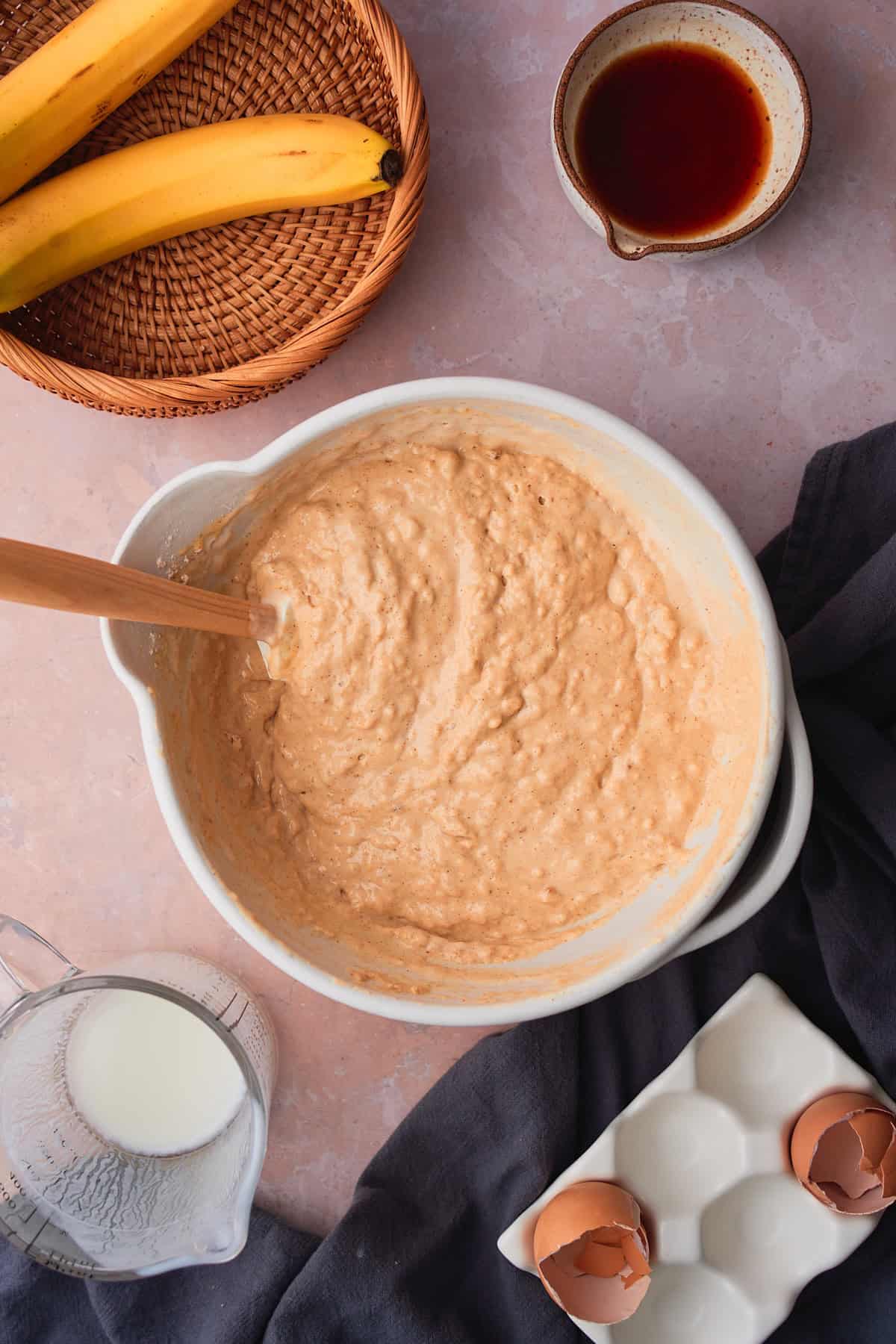 Super fluffy pancake batter in a mixing bowl being folded together with a rubber spatula surrounded by ingredients and bananas in a wicker basket. 