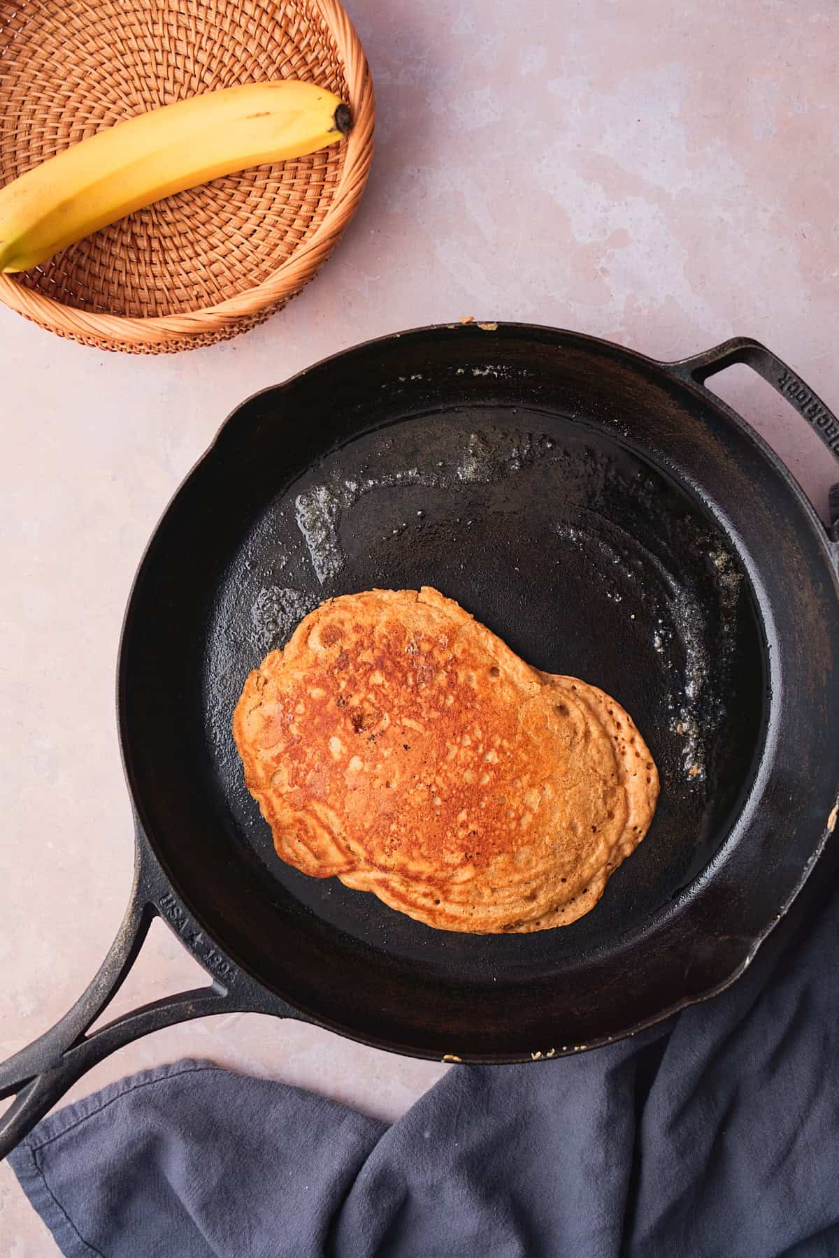 Perfectly browned and cooked pancake in a cast iron skillet with butter wit ha wicker basket nearby with a banana in the basket. 