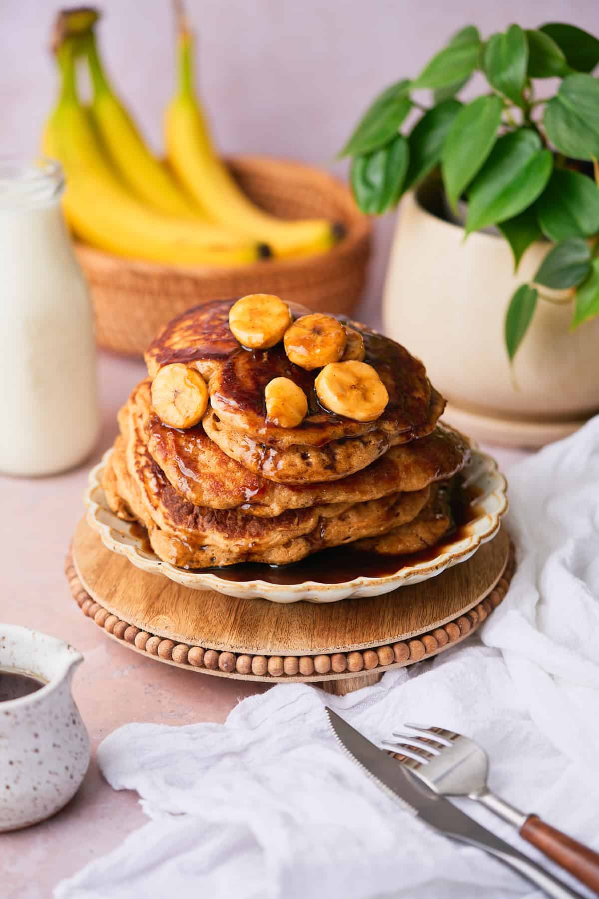Lovely stack of pancakes topped with banana slices and a brown sugar rum caramel sauce with more bananas in the background and green plant. 