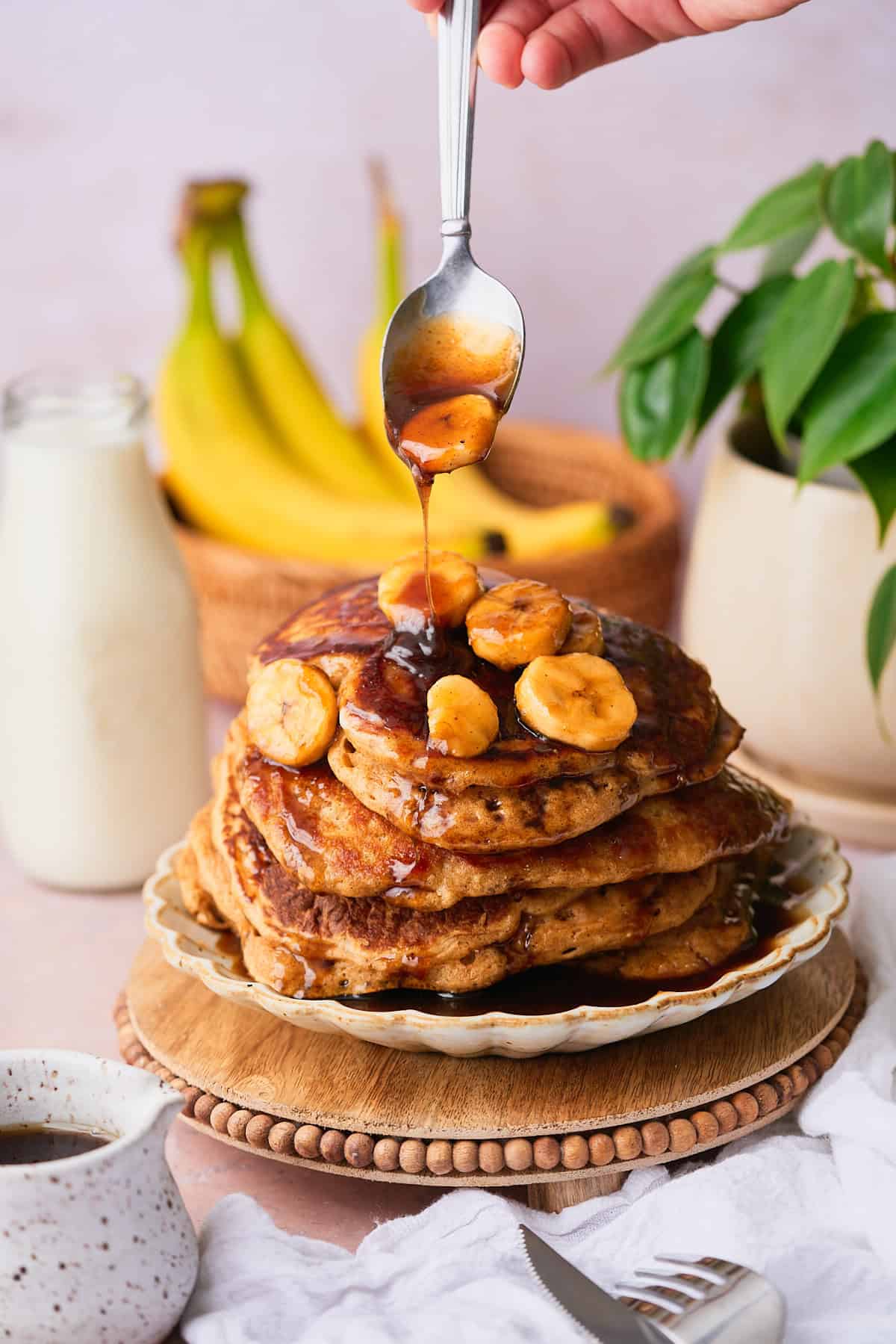 Stack of fluffy pancakes with a brown sugar bananas foster topping being dolloped onto the top, with bananas and a green plant in the background.
