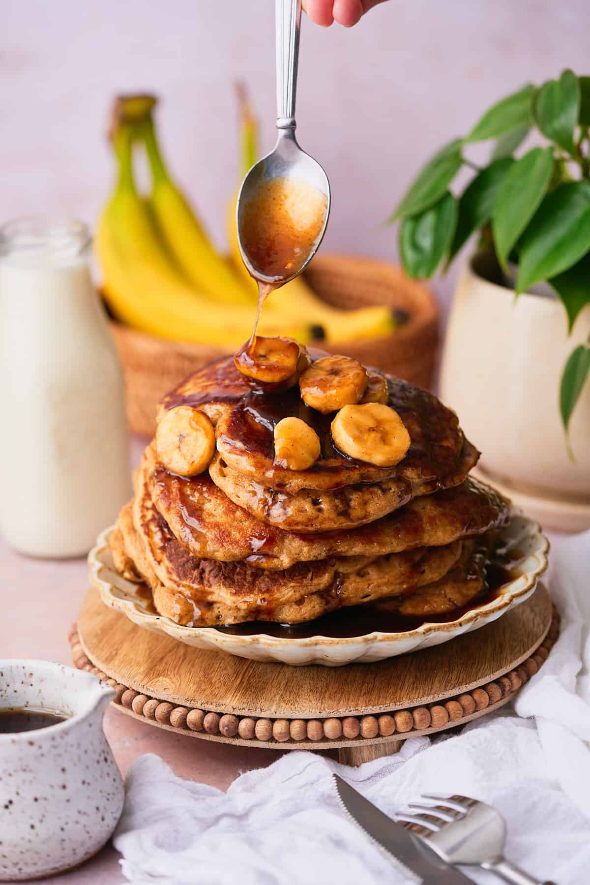 Stack of delicious pancakes with a banana topping being drizzled on top, with a glass jug of milk in the background, a green plant, and bananas in a wicker basket. 