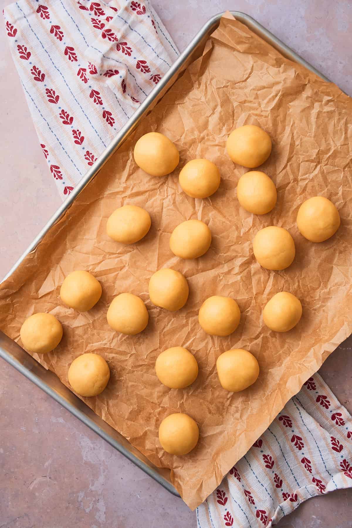 Balls of cookie dough on a parchment lined baking sheet on top of a linen napkin with heart shapes on it. 