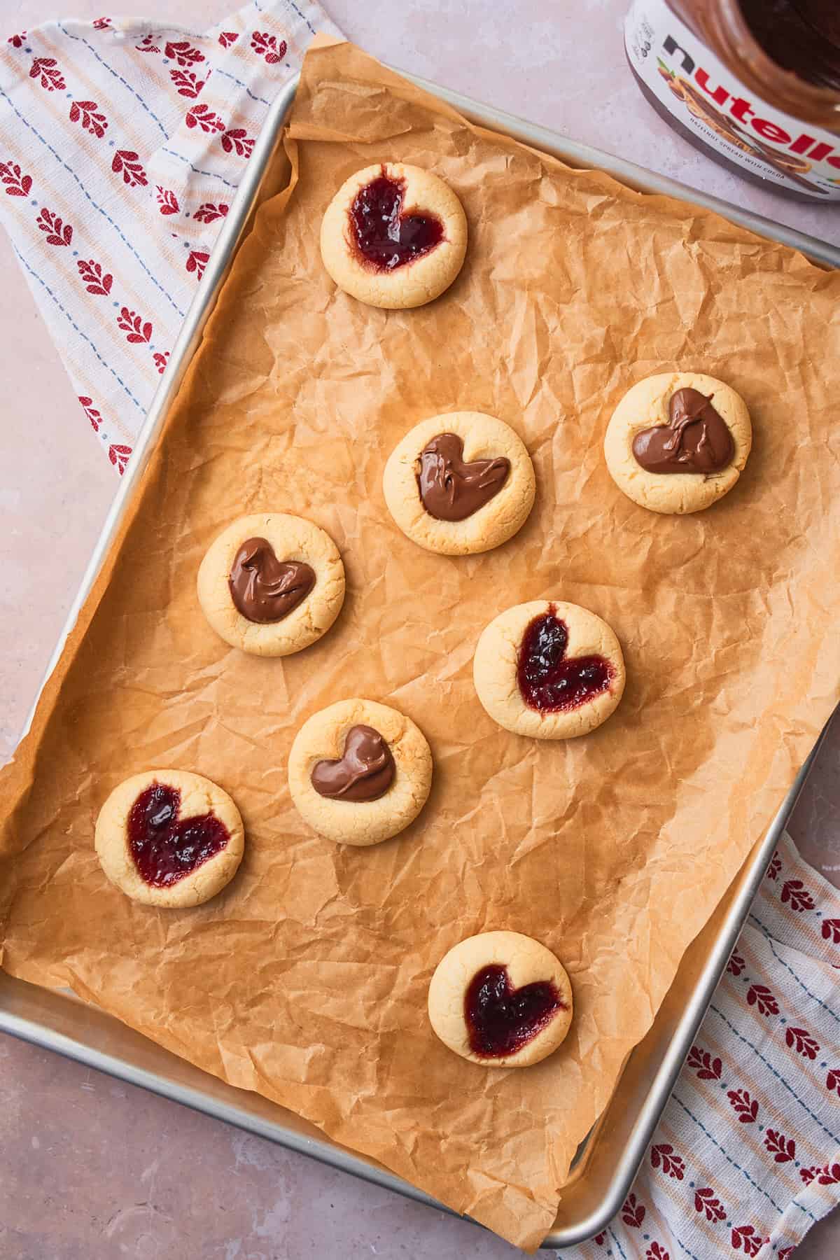 Baked cookies with a heart shape in the center, filled with Nutella and jam and cooling on a baking sheet. 
