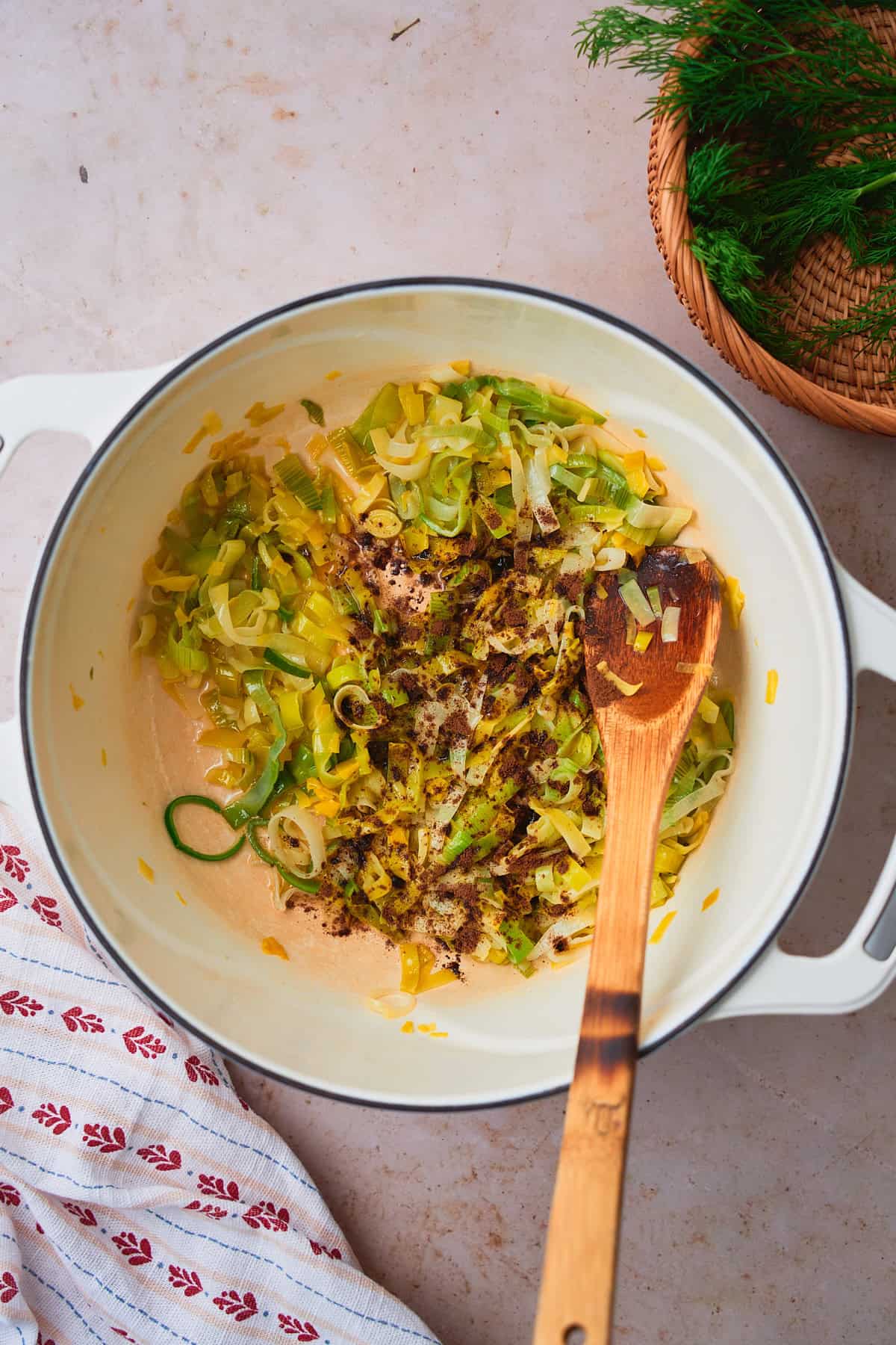 Leeks cooking in a pot with allspice added, with a wicker basket and dill inside, overtop of a cute linen napkin.