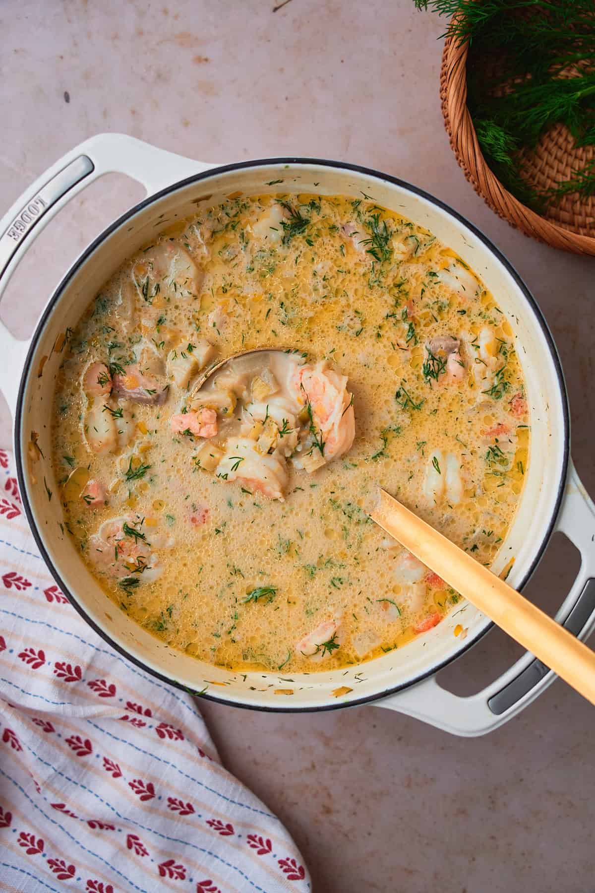Fish soup cooked to perfection with lots of fresh dill added to the creamy broth in a white Dutch oven, surrounded by a wicker basket filled with fresh dill, on top of a linen napkin with little red floral print. 