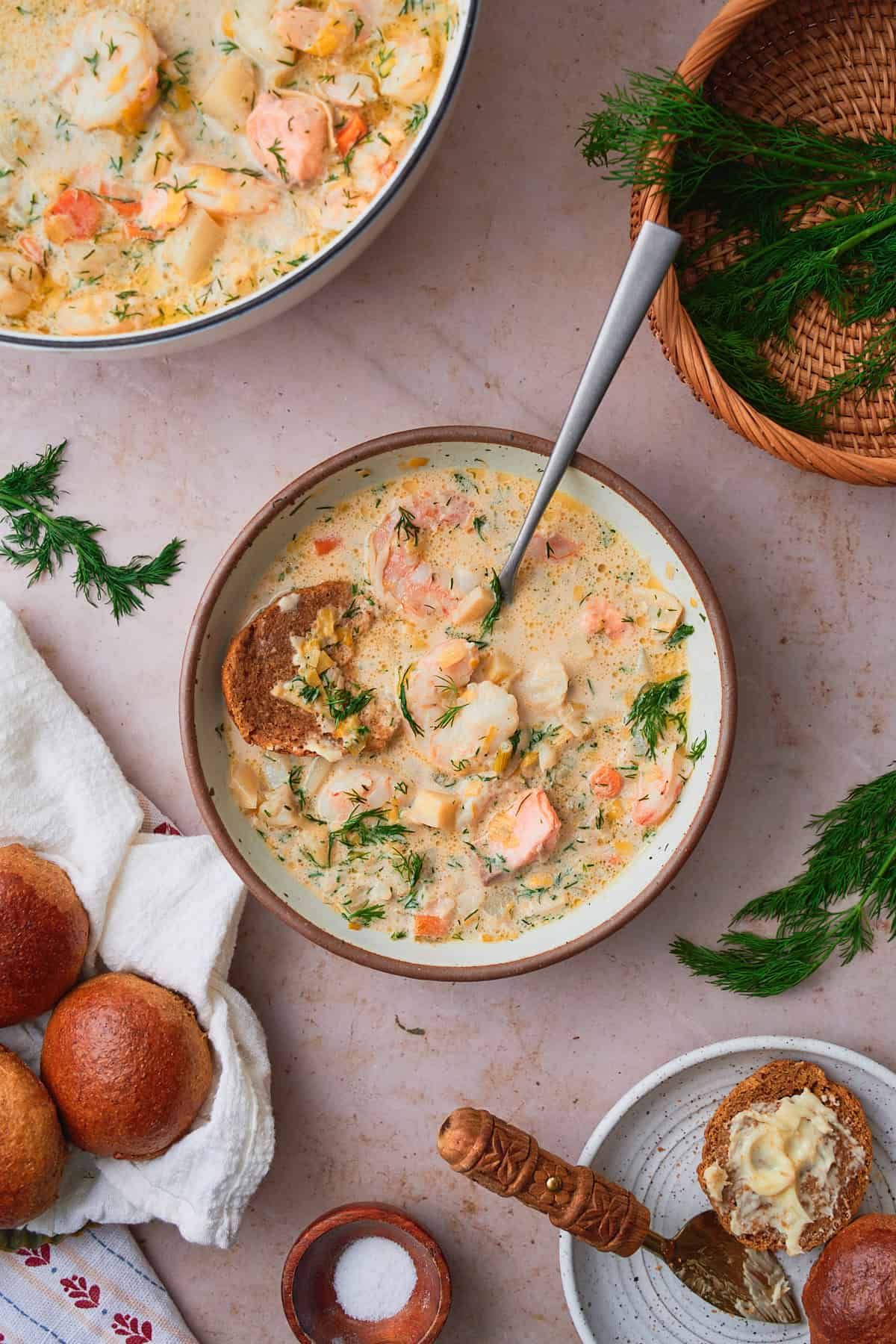 Overhead shot of a bowl of Norwegian fish soup surrounded by fresh dill, rye bread rolls, and butter. 