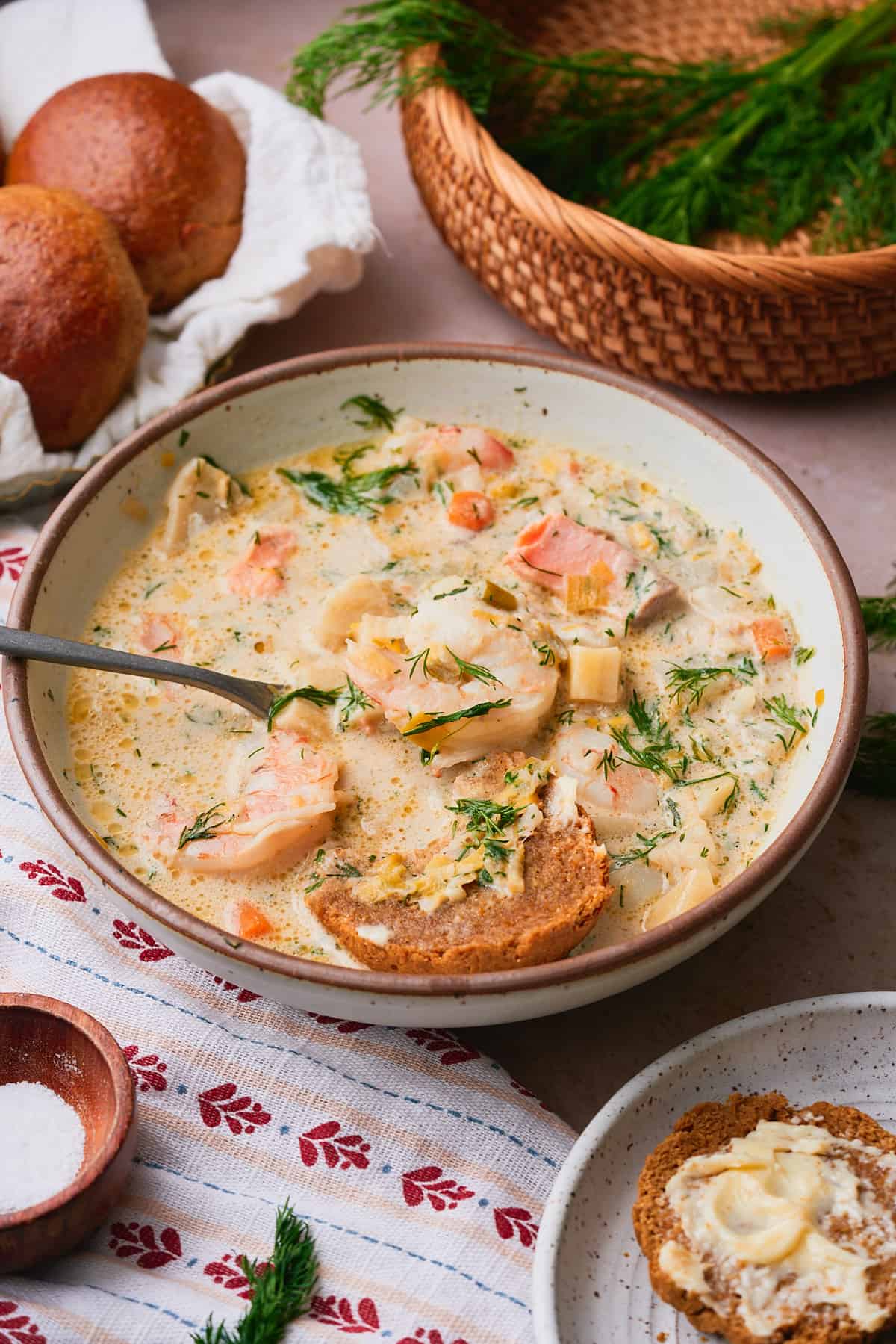 Bowl of creamy Norwegian fish soup with a cut rye roll in the bow, surrounded by a wicker basket with fresh dill in the basket, an a cute Nordic napkin in the foreground.
