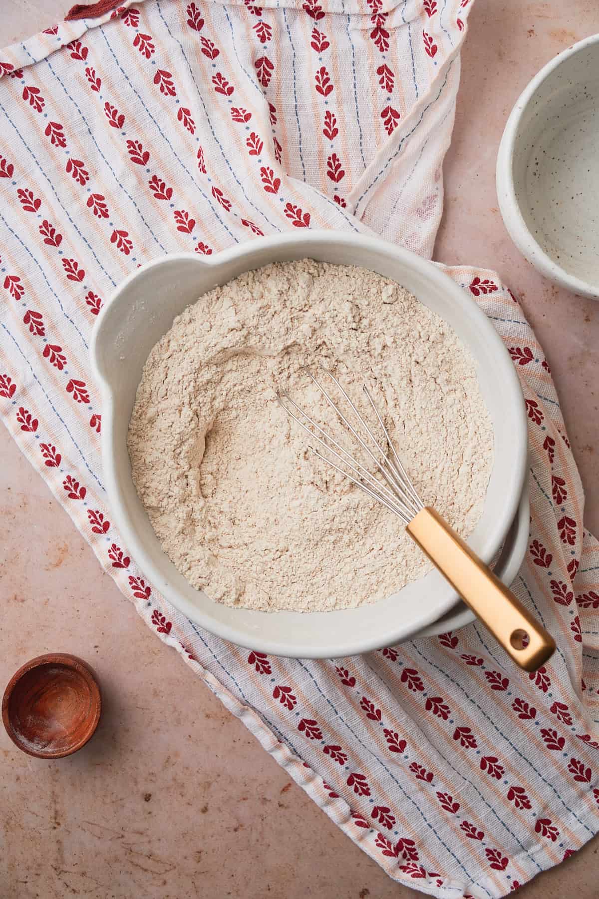 Flours and salt mixed together in a bowl with a whisk on top of a linen napkin with a cute heart print.