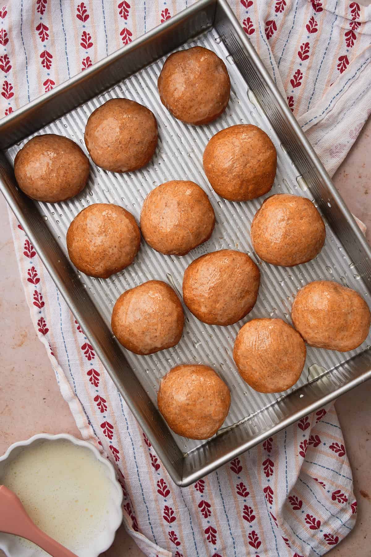 Glossy rye buns in a metal baking sheet getting ready to go in the oven.