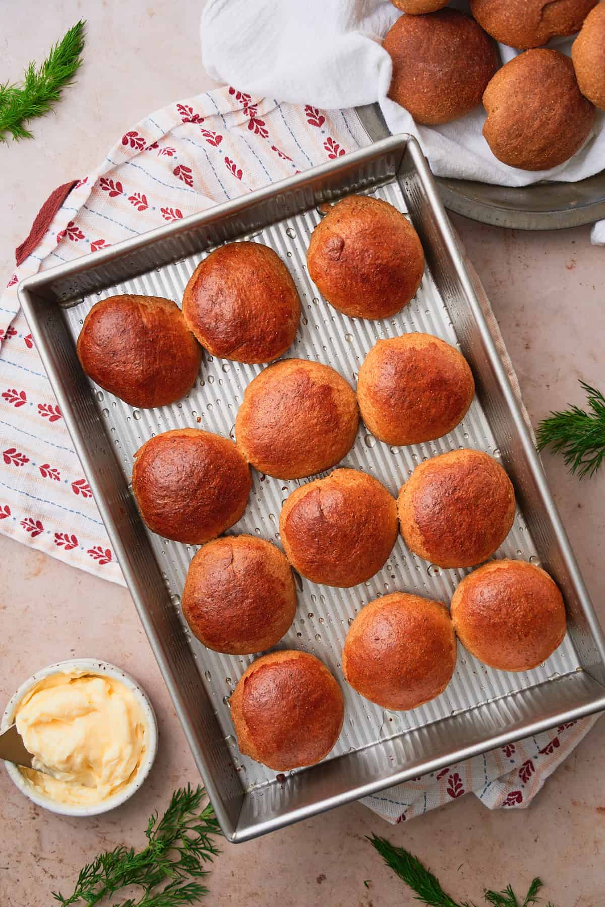 Baked and golden brown rye rolls in a metal pan surrounded by butter, fresh dill, and plates with more buns on them.