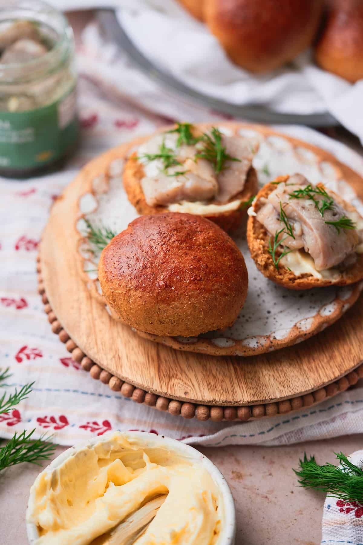 Soft and fluffy rye roll on a ceramic plate on top of a wooden board with a patterned linen napkin underneath, with halved pieces of bread topped with pickled herring and fresh dill.