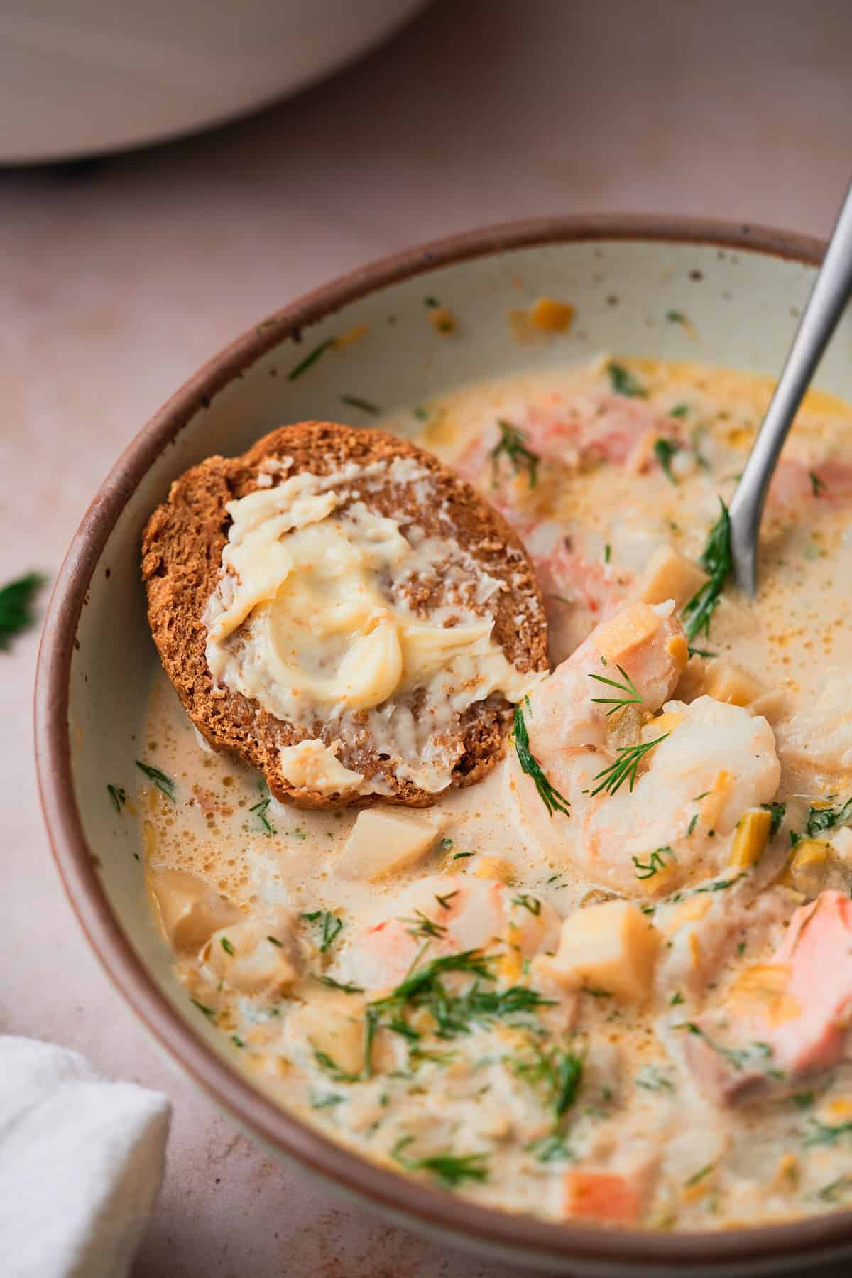 Close up shot of butter smeared on a rye dinner roll cut in half in a bowl of creamy fish soup.