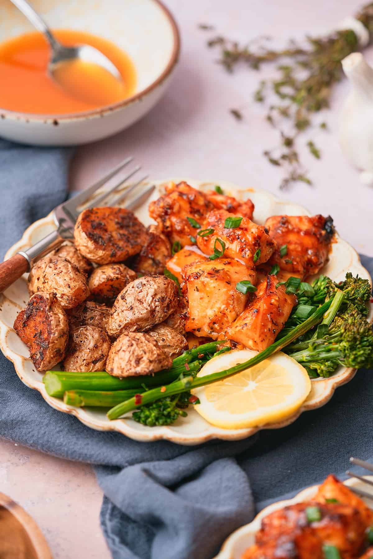 Salmon bites in a sticky sweet sauce on a ruffled plate served with potatoes, broccolini and lemon.