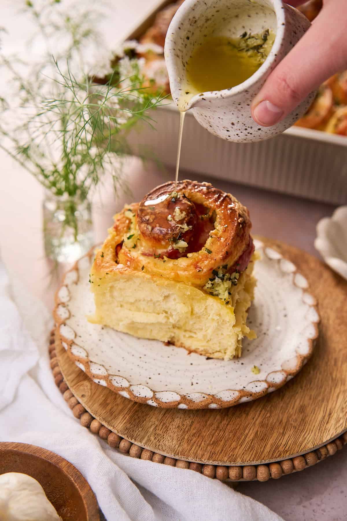 Hand drizzling a fluffy ham and cheese roll with garlic butter on a ruffled white ceramic plate on top of a wooden charger plate, with green and white flowers in the background. 