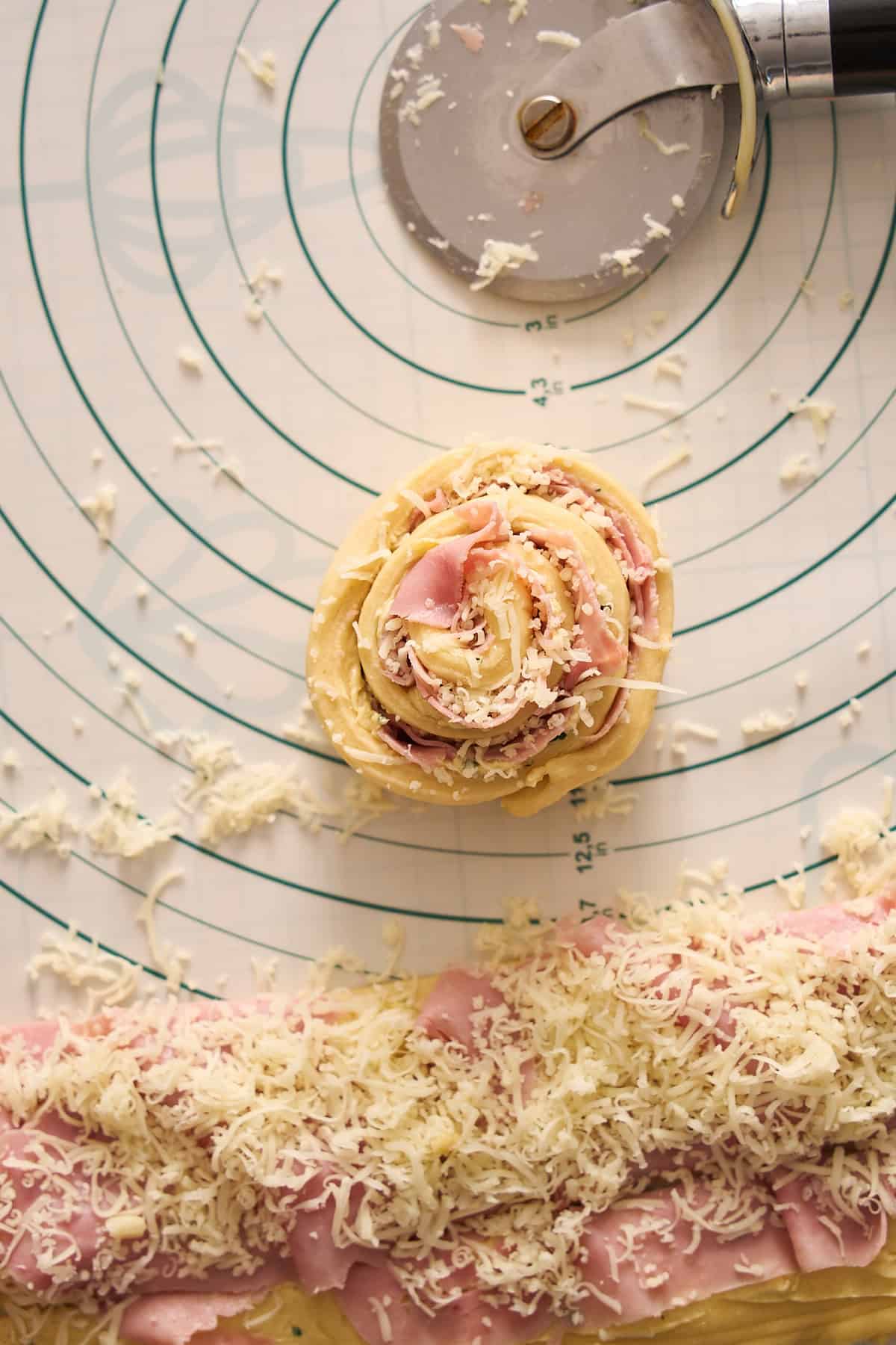 Tightly rolled ham and cheese roll on a silicone baking mat with a pizza cutter nearby. 