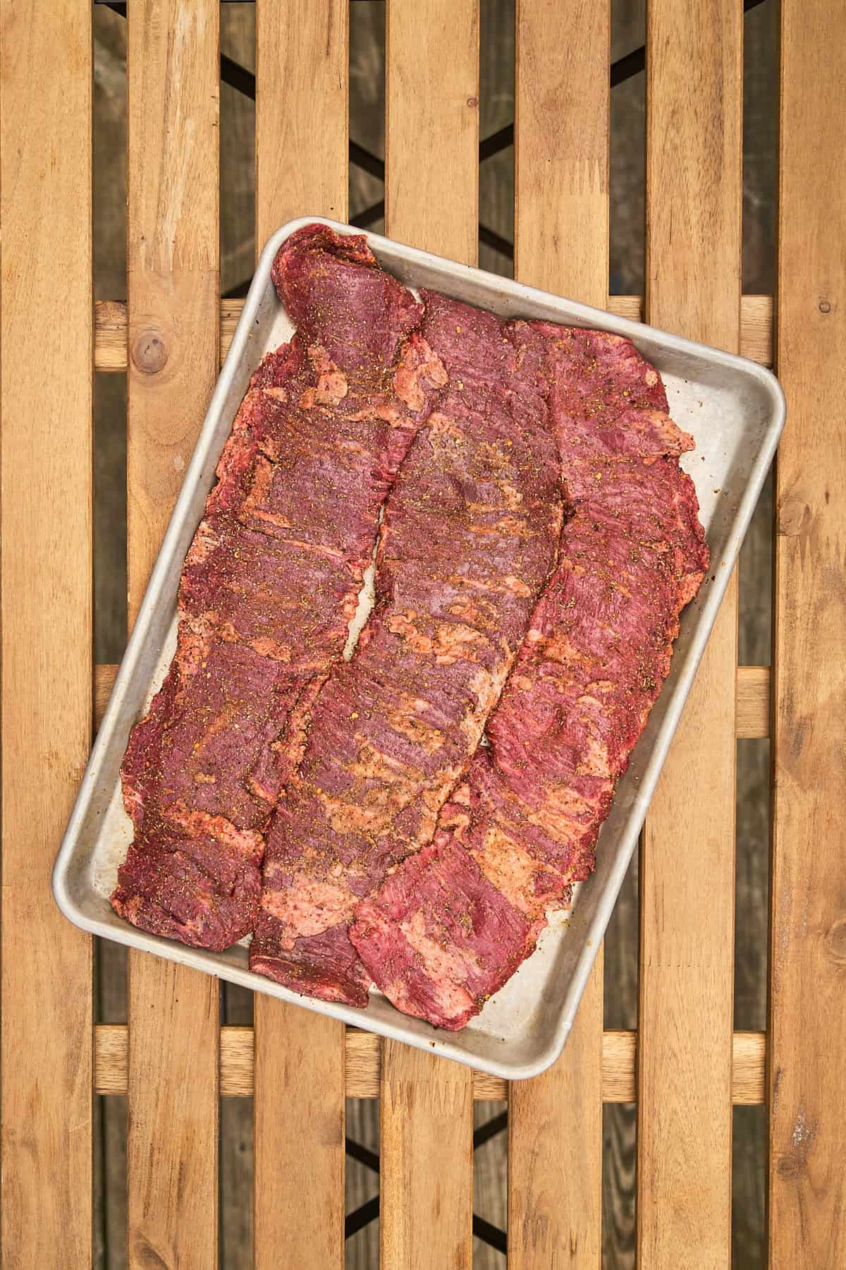 Seasoned skirt steaks on a silver baking sheet on a wooden table.