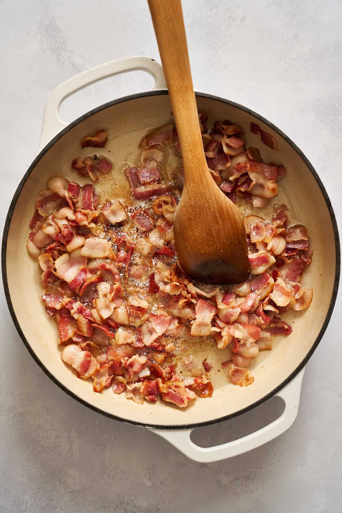 Bacon pieces cooking in a large skillet with a wooden spoon stirring them.