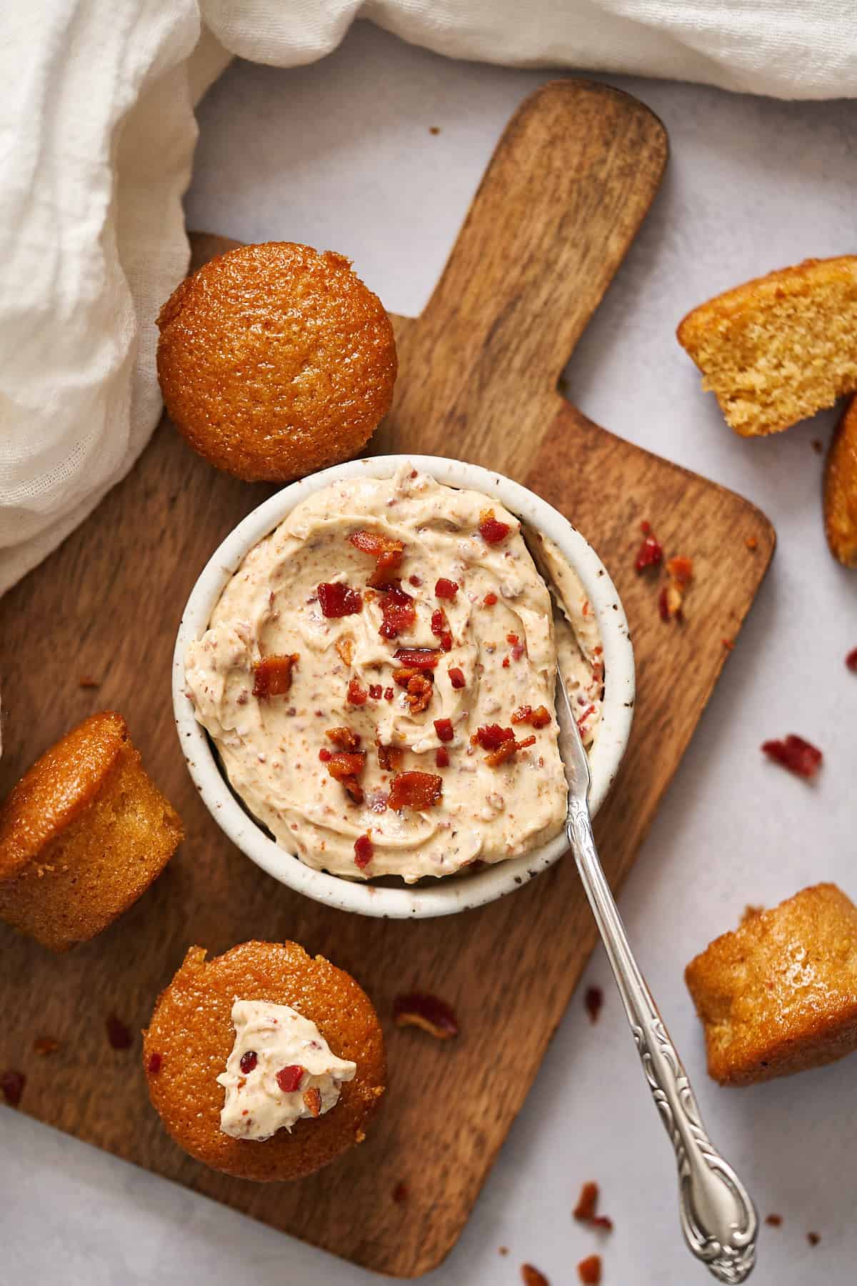 Overhead view of bacon butter in a ramekin with a butter knife in it, surrounded by small cornbread muffins.