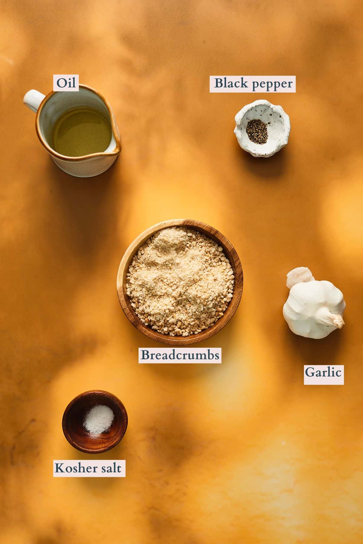 Ingredients to make a garlic breadcrumb topping laid out on a bright yellow backdrop and labeled to denote each ingredient.