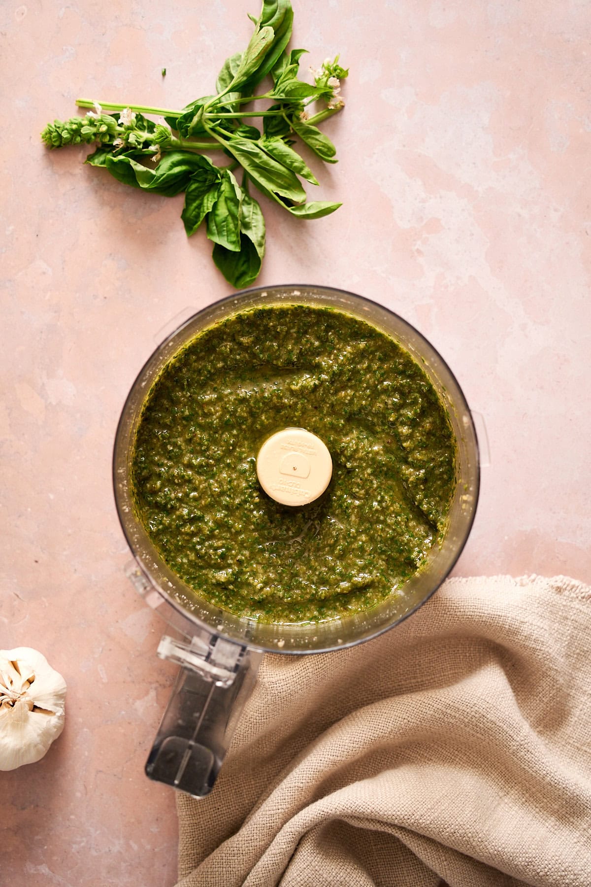 Smooth and creamy basil pesto in a food processor surrounded by garlic, a linen napkin, and ggresh basil.