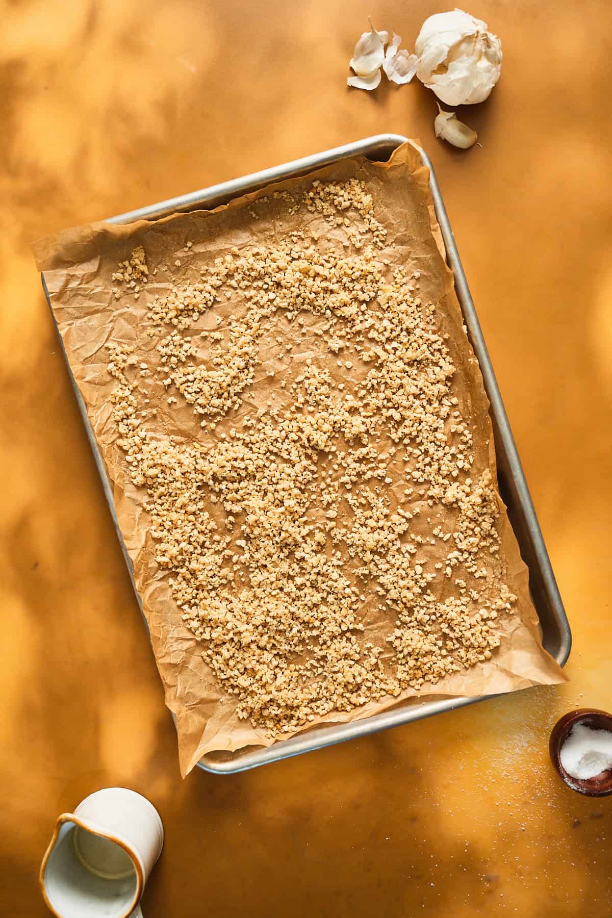 Breadcrumbs on a baking sheet seasoned with salt, pepper, grated garlic, and oil.
