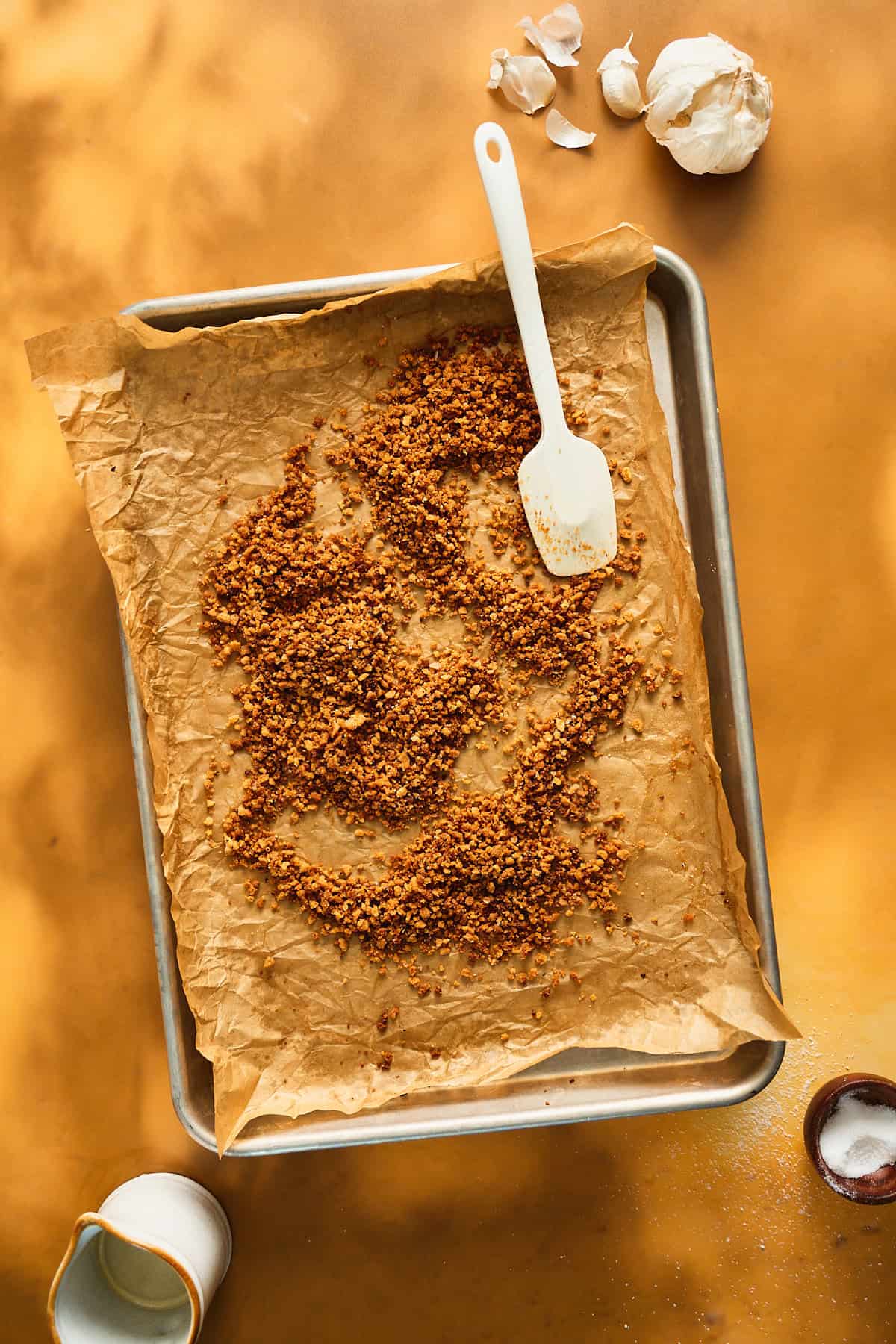 Breadcrumbs that have browned deeply on a baking sheet on a yellow backdrop.