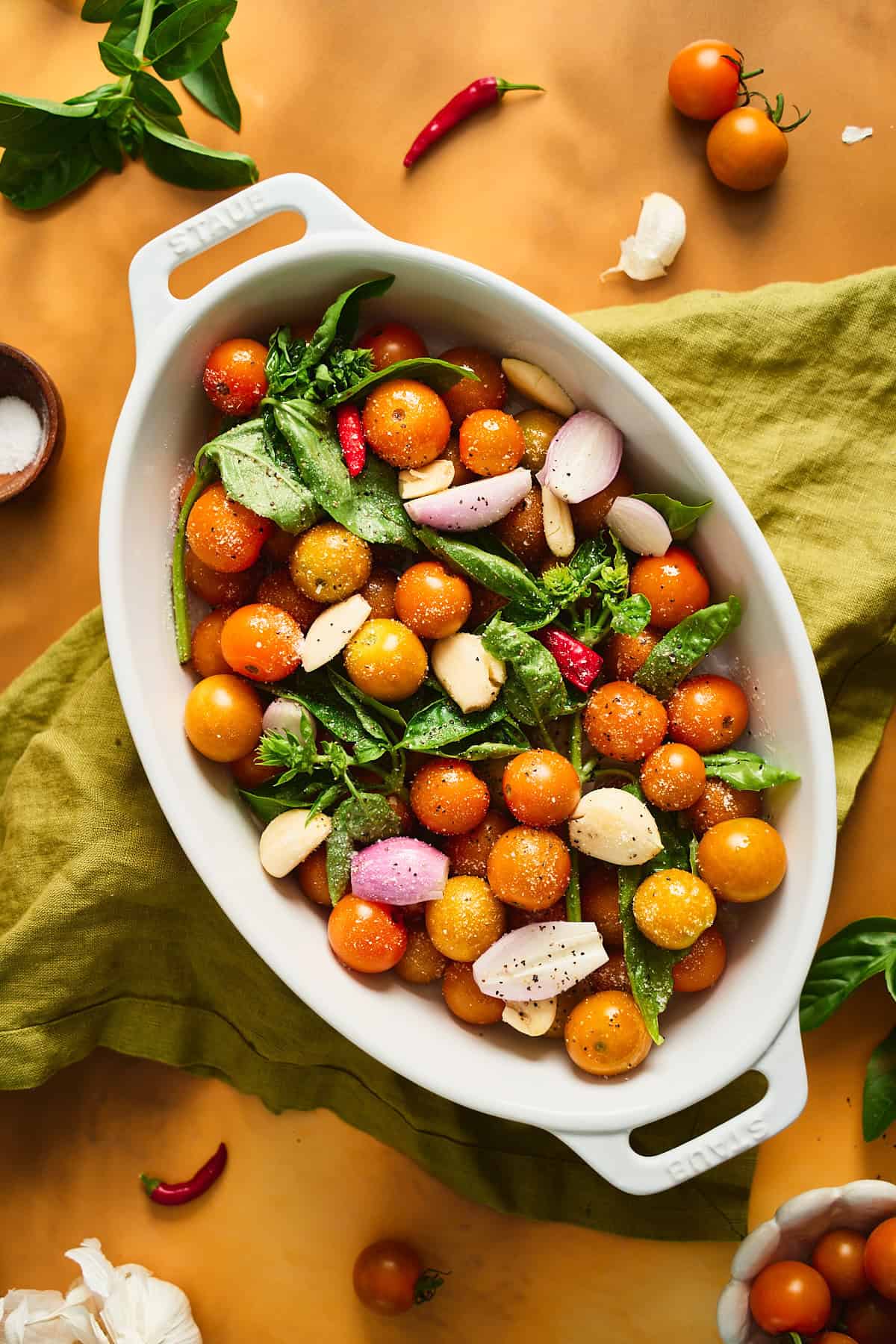 Shallow baking dish with vibrant colors of Sungold tomatoes, garlic, shallots, chilis, and basil in a white baking dish getting ready to go into the oven.