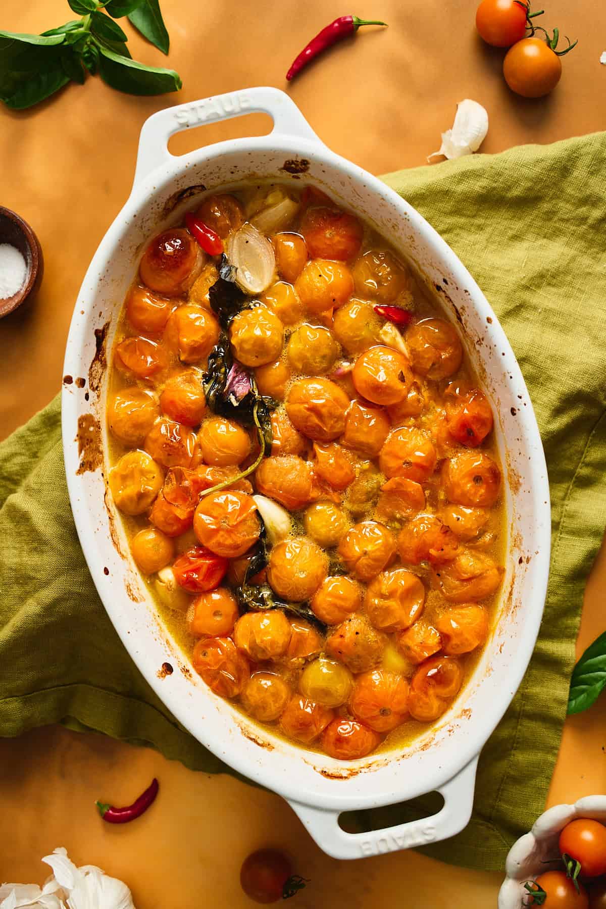 Roasted bright orange cherry tomatoes alongside garlic and shallots on a deep yellow backdrop with a green linen underneath.