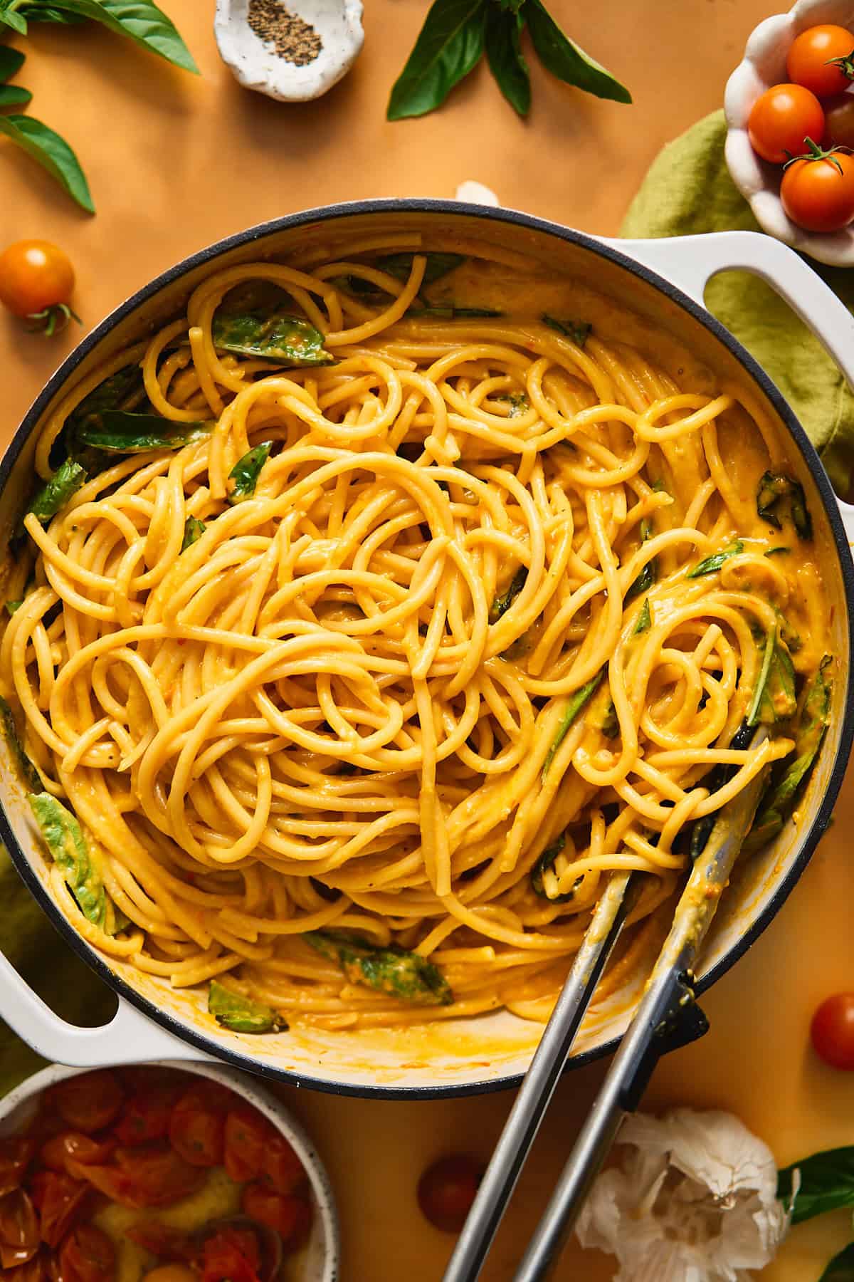Bucatini noodles in a large pan coated with a golden creamy sauce, with tongs in the pan.