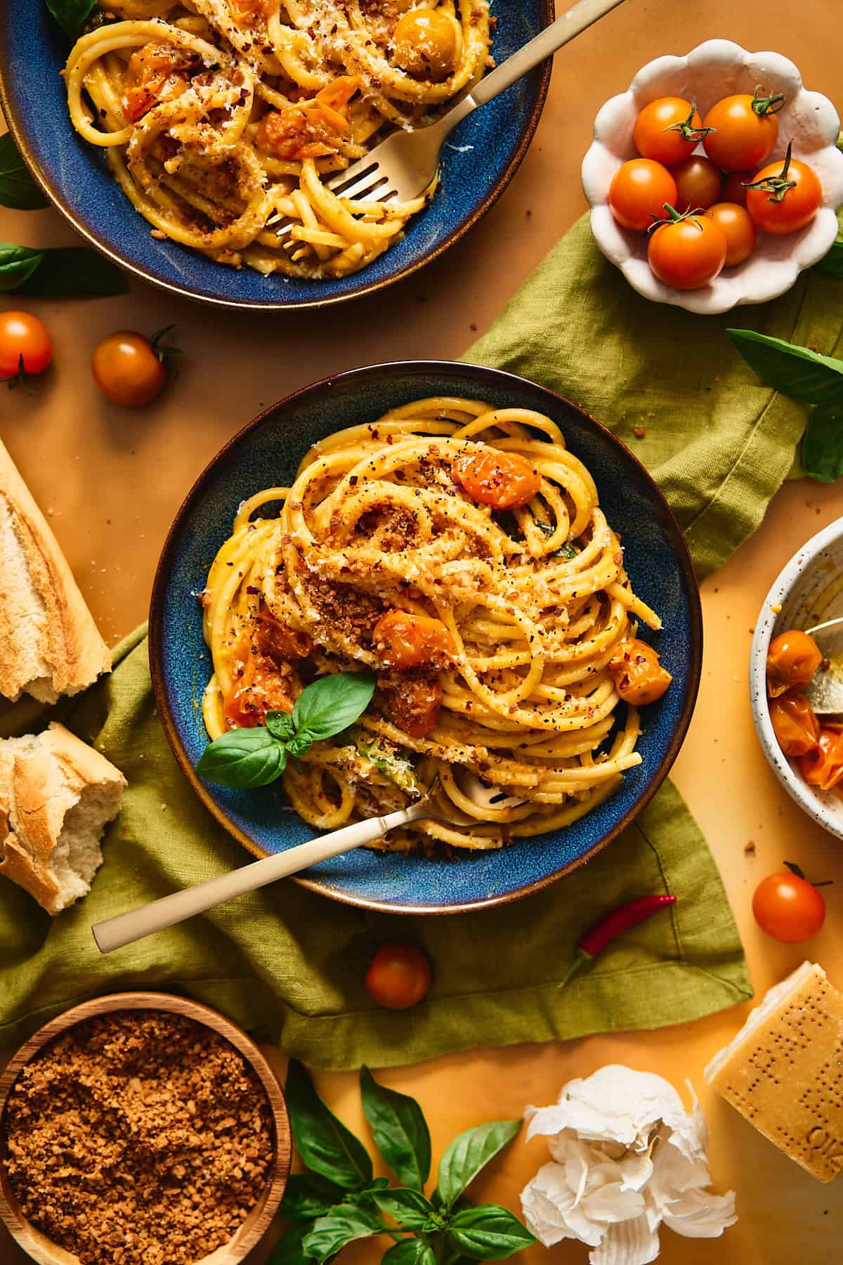 Luscious orange Sungold pasta in a blue bowl, topped with burst orange cherry tomatoes, basil, breadcrumbs, and surrounded by ingredients to make the pasta.