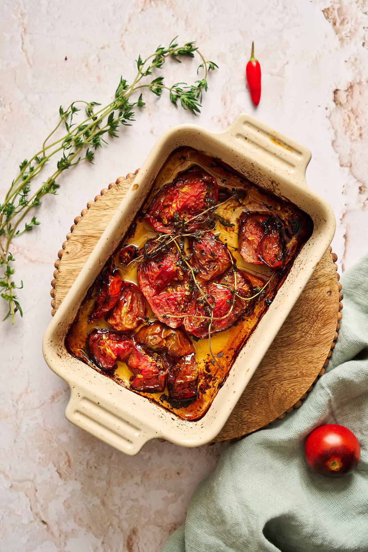 Roasted tomatoes in a casserole dish surrounded by a light blue linen, more fresh tomatoes and herbs.