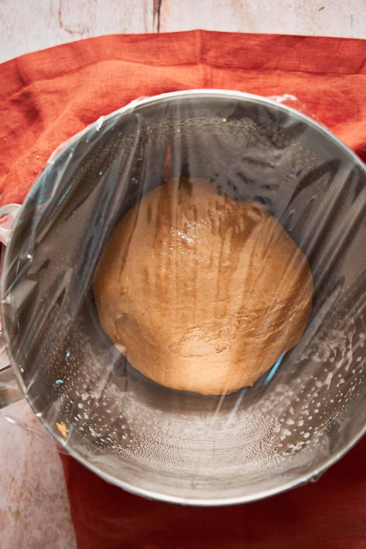 Cinnamon roll dough covered with plastic wrap in a metal bowl.