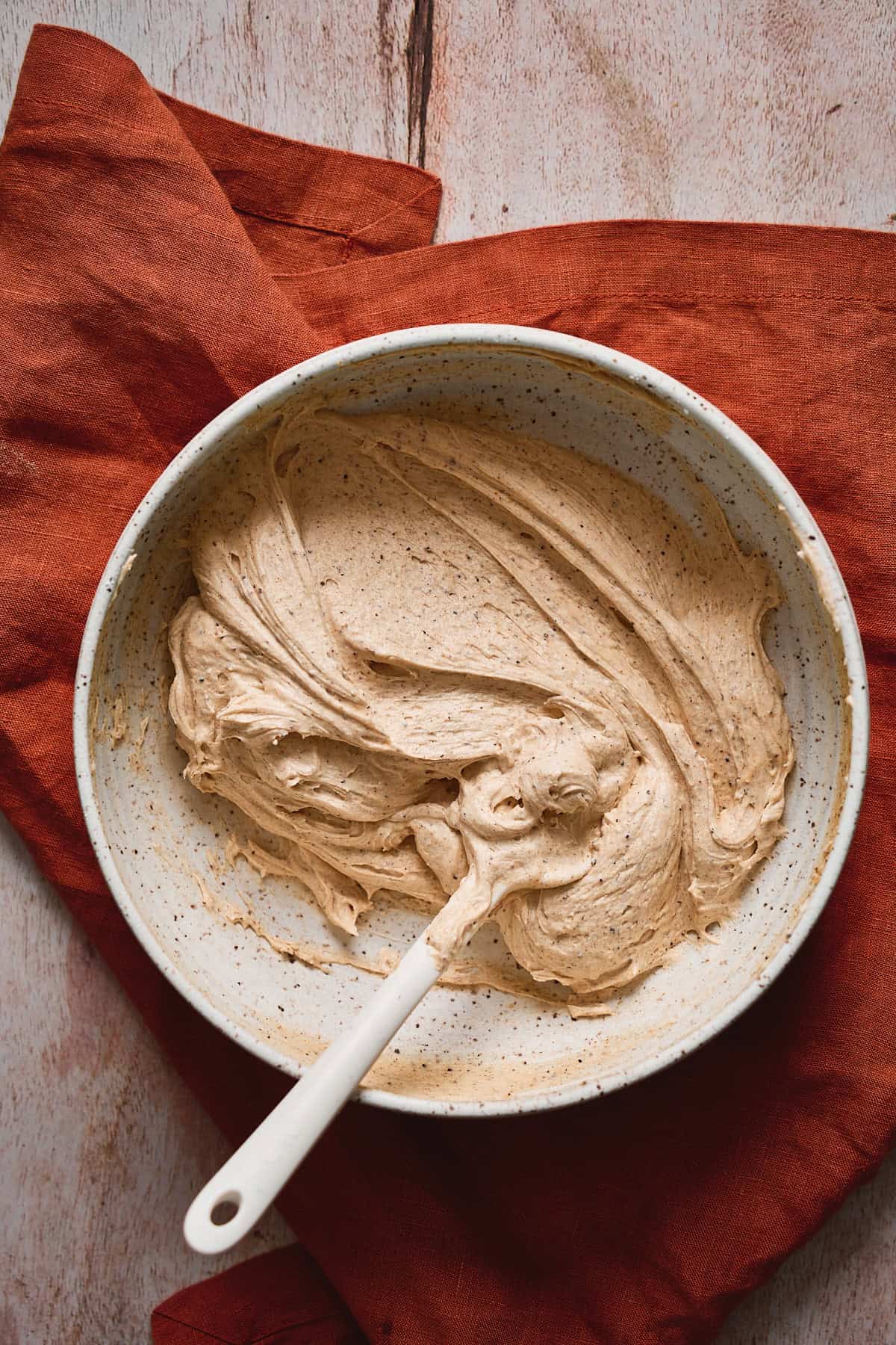 Cream cheese frosting mixed in a bowl with a rubber spatula on a terra cotta linen.