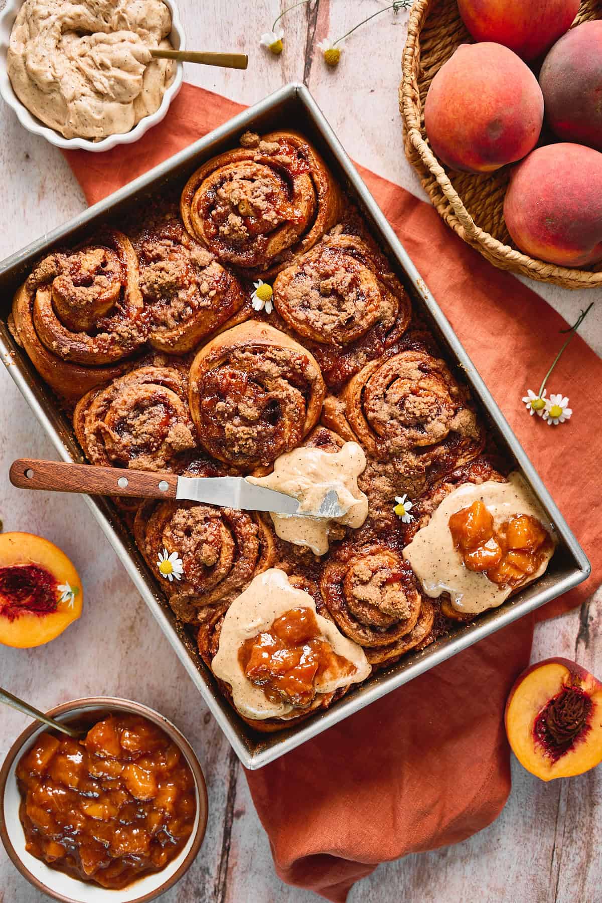 Tray of peach cinnamon rolls topped with cream cheese frosting that is melting on it from the warmth of the cinnamon rolls, with a peach topping on top of the rolls, with peaches and chamomile flowers on the scene.