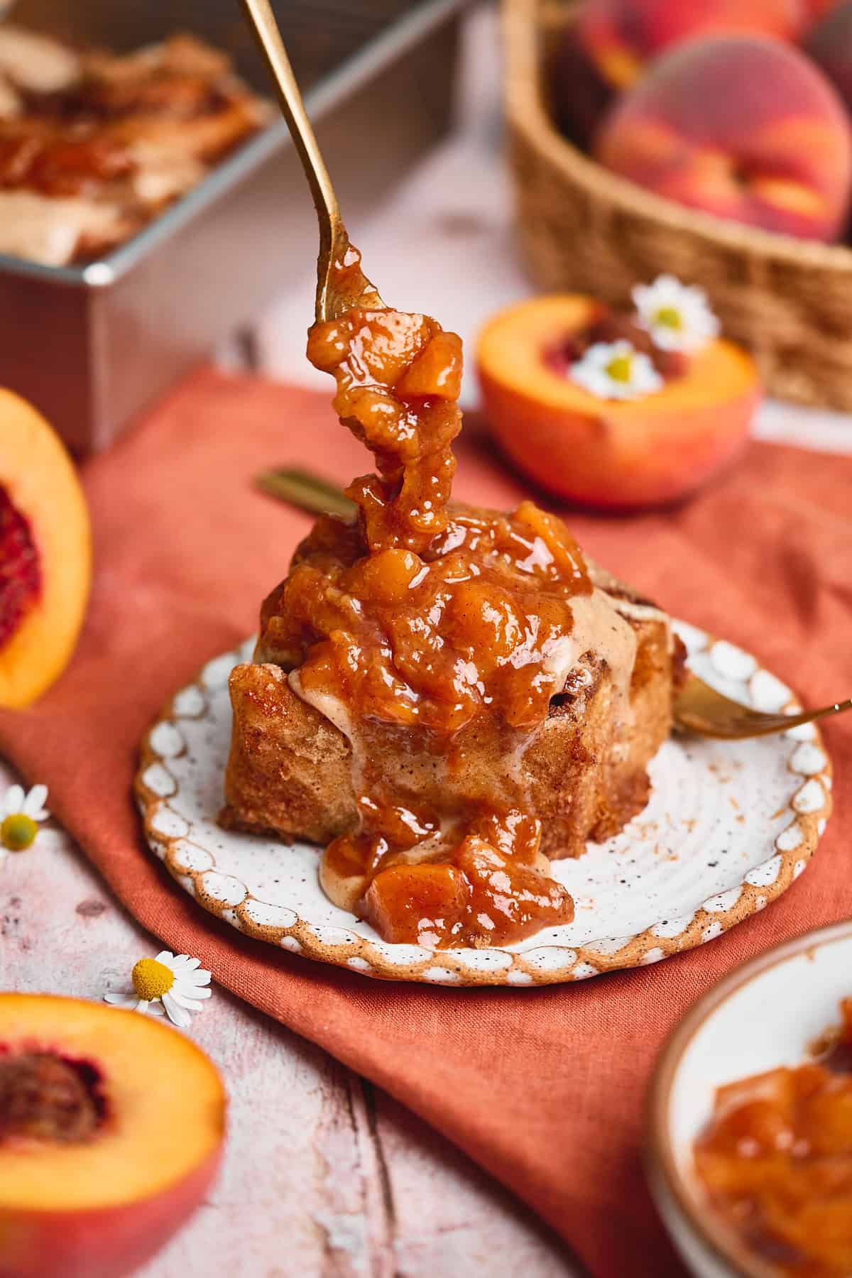 Beautiful peach cobbler cinnamon roll on a ruffled ceramic plate topped with a peach jam and cream cheese frosting on top of a terra cotta linen napkin surrounded by peaches and small chamomile flowers.