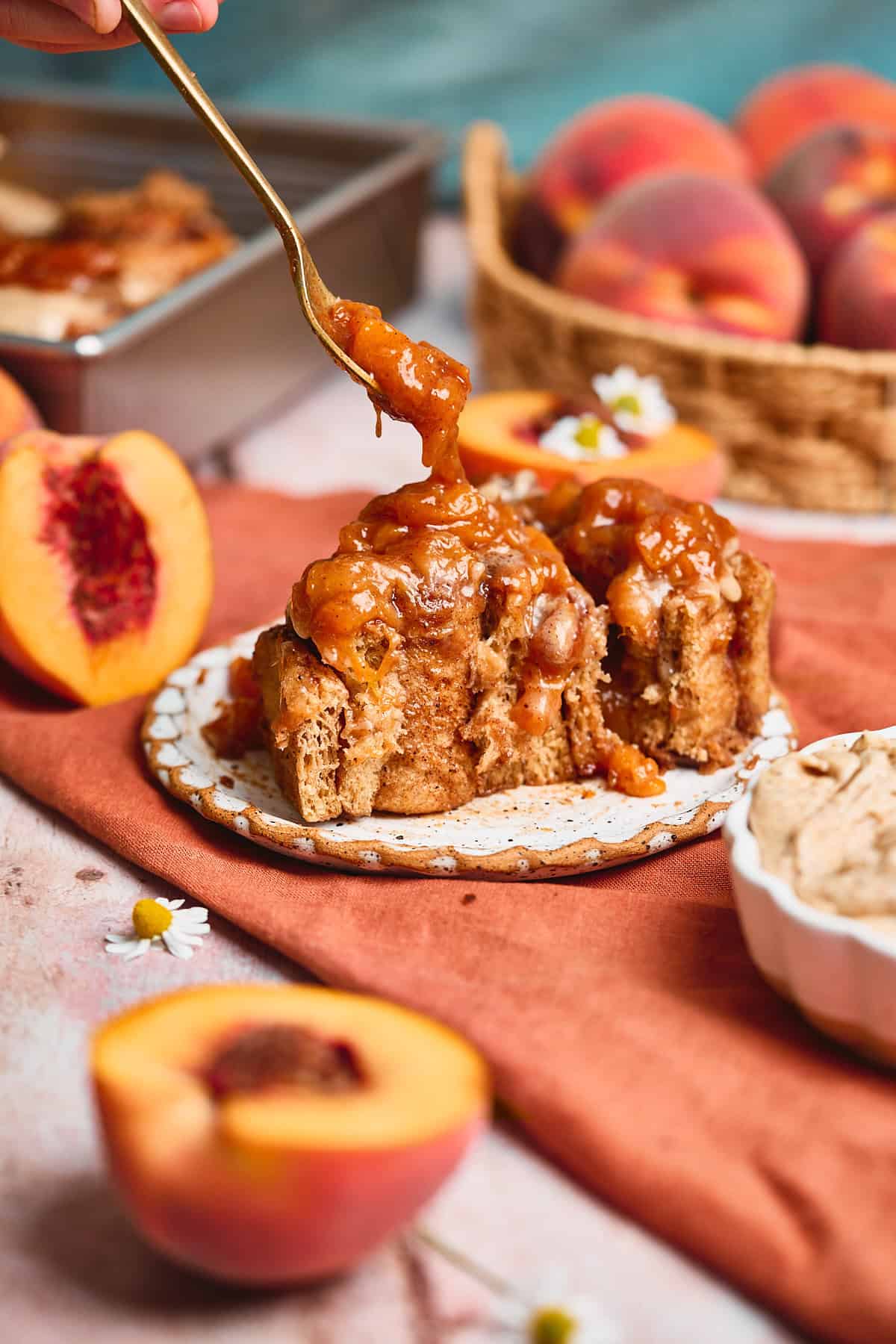 Cinnamon roll that has been cut in half, with peach jam being spooned on top of the roll, surrounded by peaches and small chamomile flowers.