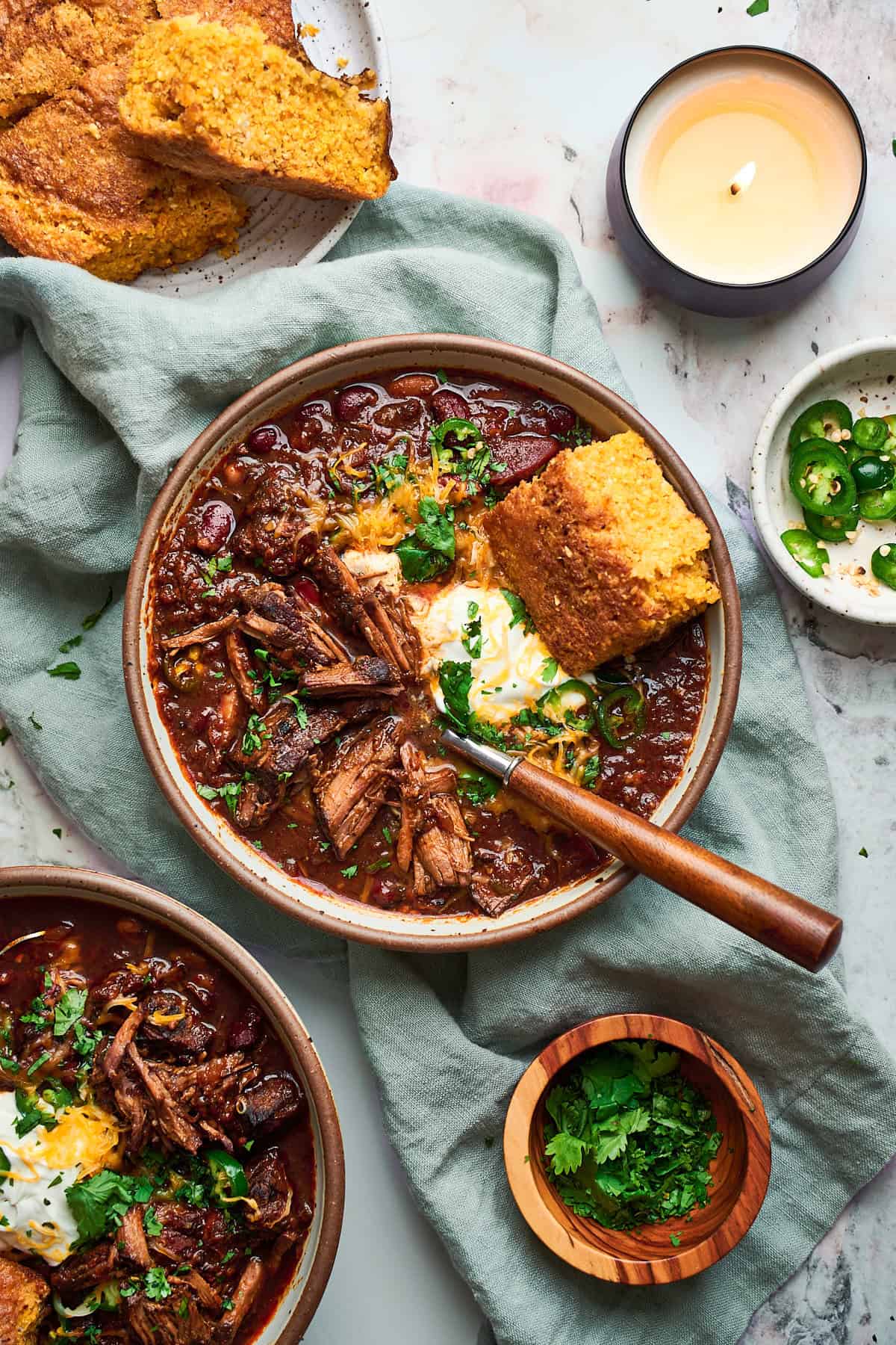 Flat lay shot of a large bowl of chuck roast chili topped with sour cream, cheese, cornbread, and jalapeno pieces surrounded by a candle and more bowls of chili and plates of cornbread.