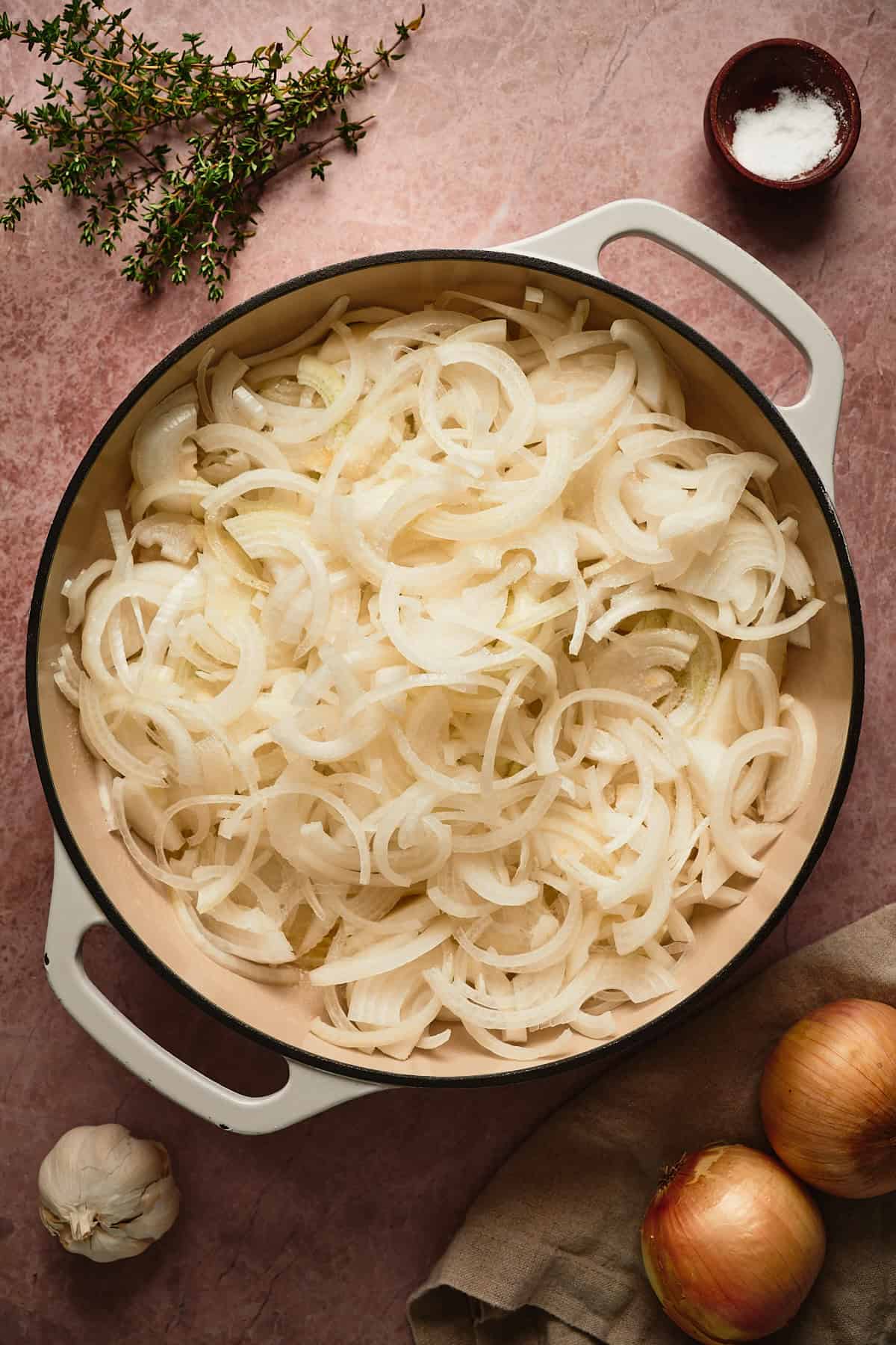 Yellow onions in a large skillet surrounded by fresh thyme, garlic, more onions, and salt. 