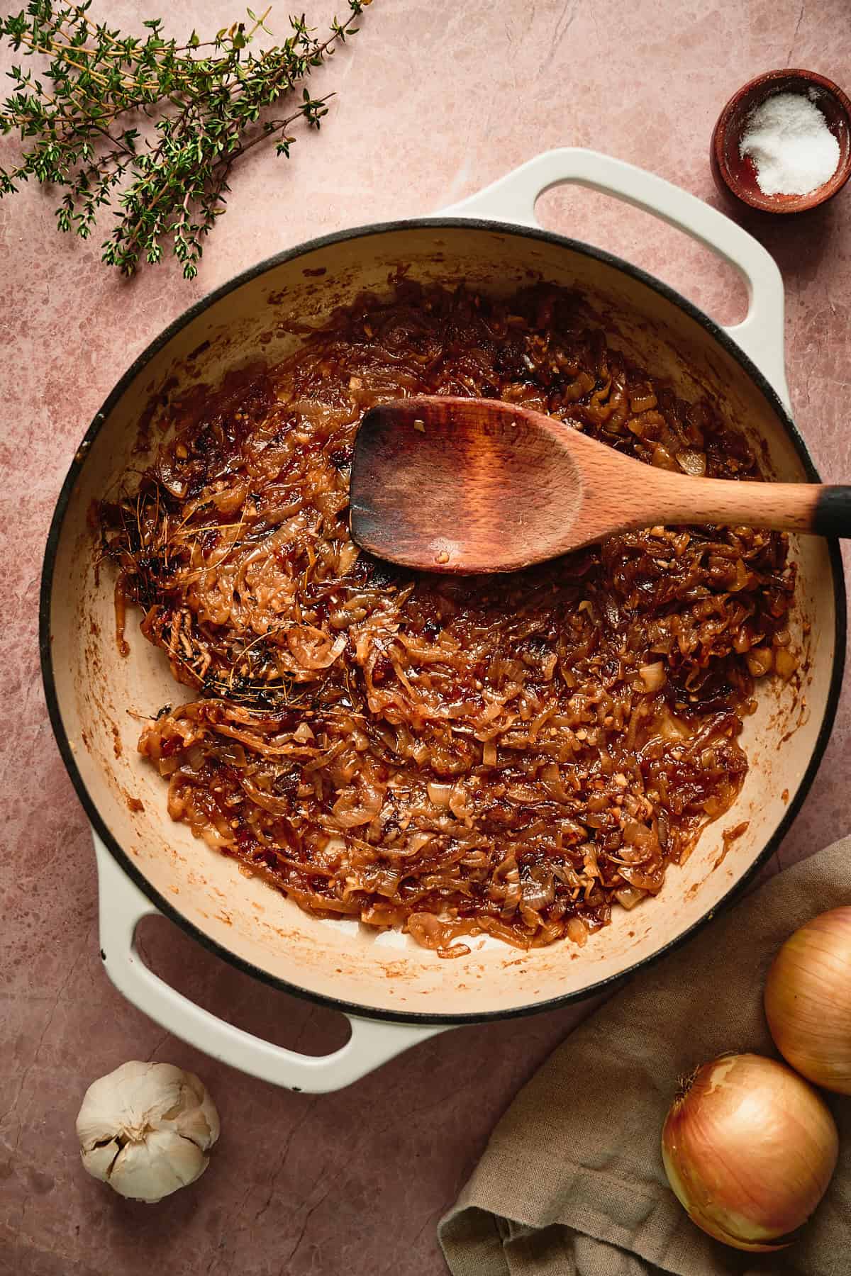 Cooked and deeply caramelized onions in a skillet surrounded by fresh thyme, garlic, more onions, and salt.