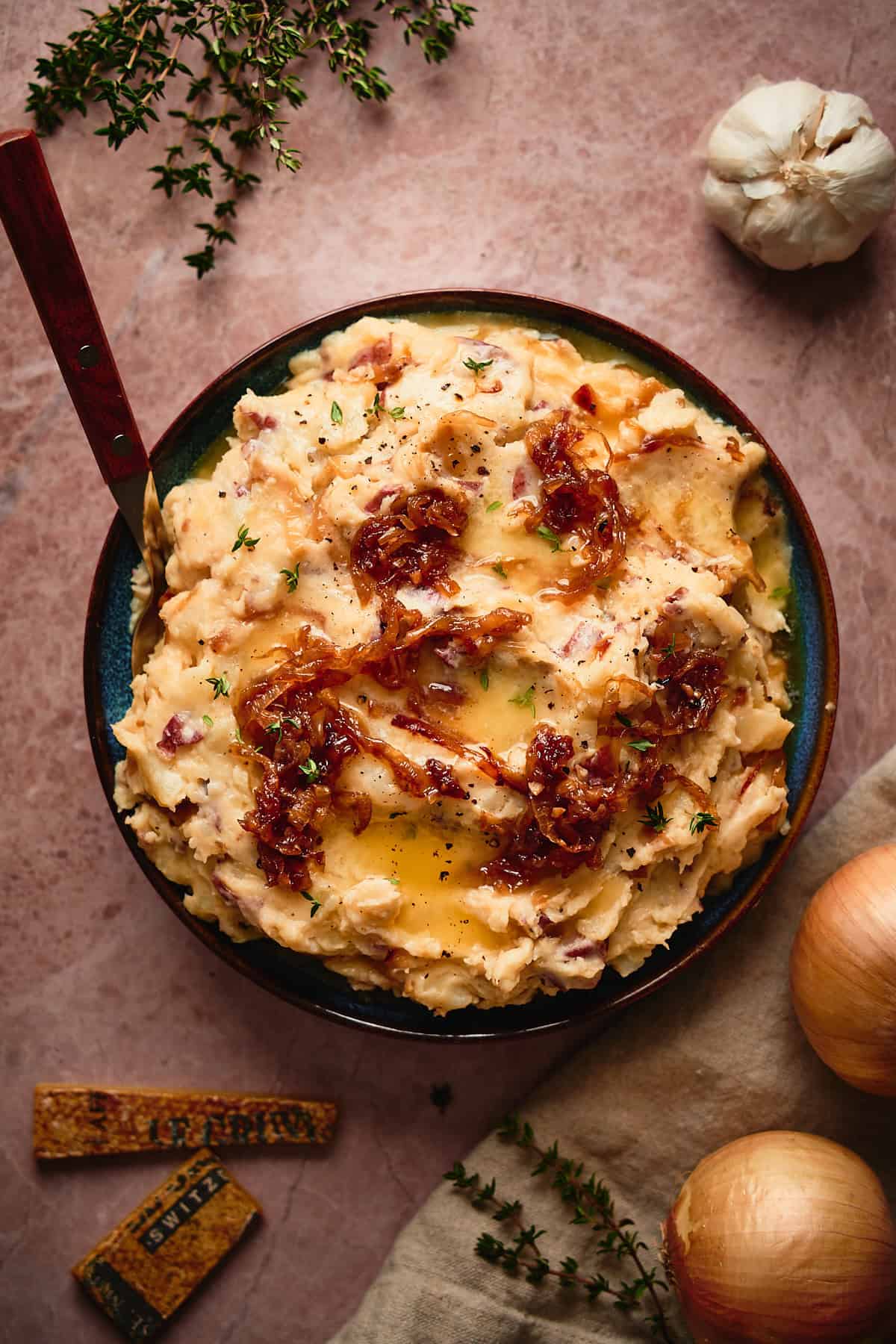 Shot of a beautiful bowl of French onion mashed potatoes on a warm colored marble backdrop, with cheese rinds, onions, thyme sprigs, and garlic surrounding the bowl. 