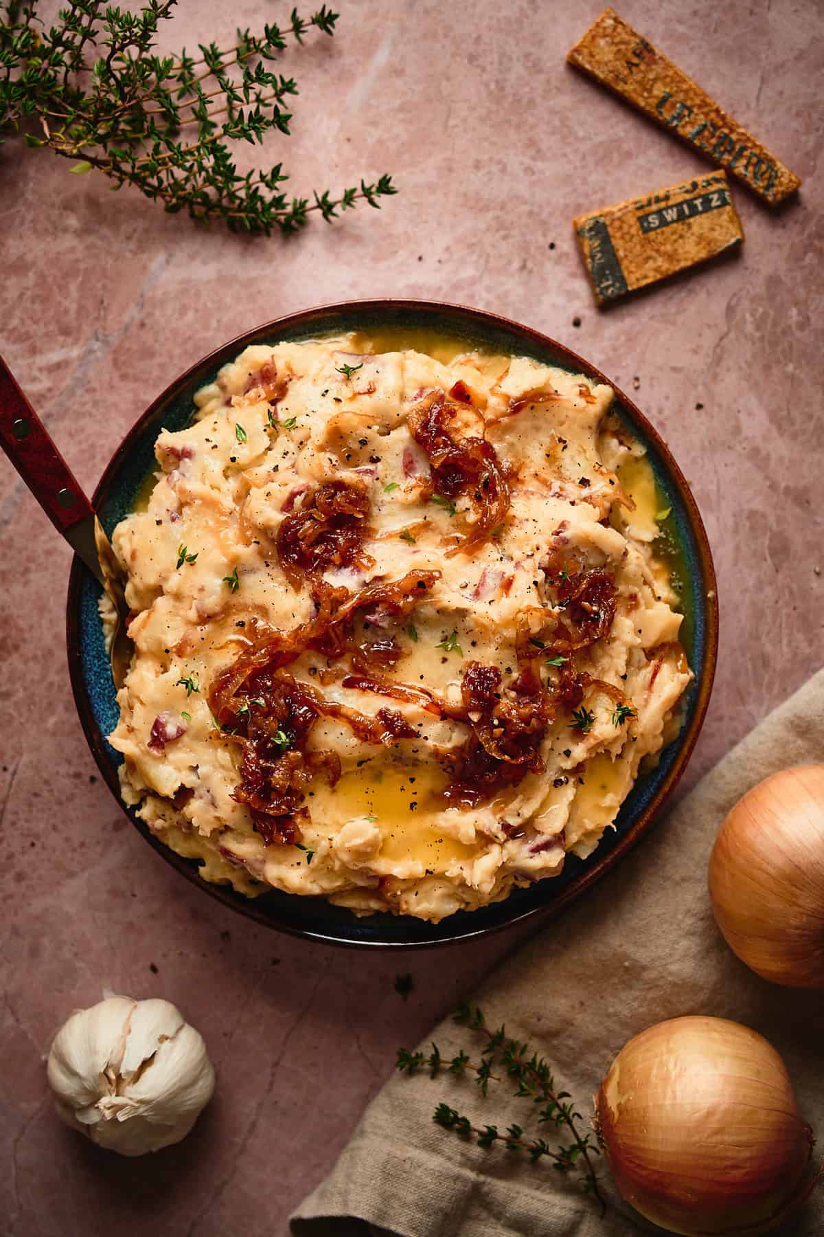Bowl of super creamy and buttery french onion mashed potatoes topped with caramelized onions, surrounded by more fresh onions, thyme, and cheese rinds.