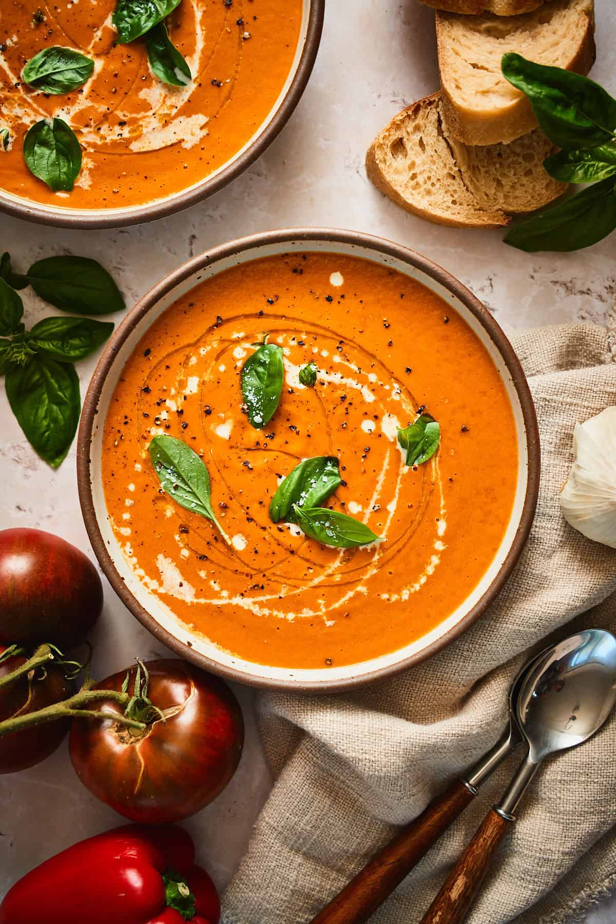stunning bowl of creamy tomato and red pepper soup topped with basil, cream, pepper, and oil, surrounded by garden fresh tomatoes, peppers and more.